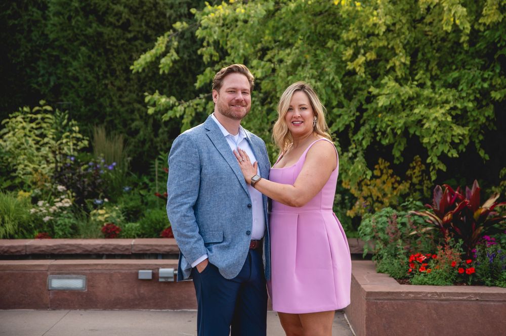 A couple poses together in a garden setting, with the woman wearing a pink dress and the man in a blue blazer.