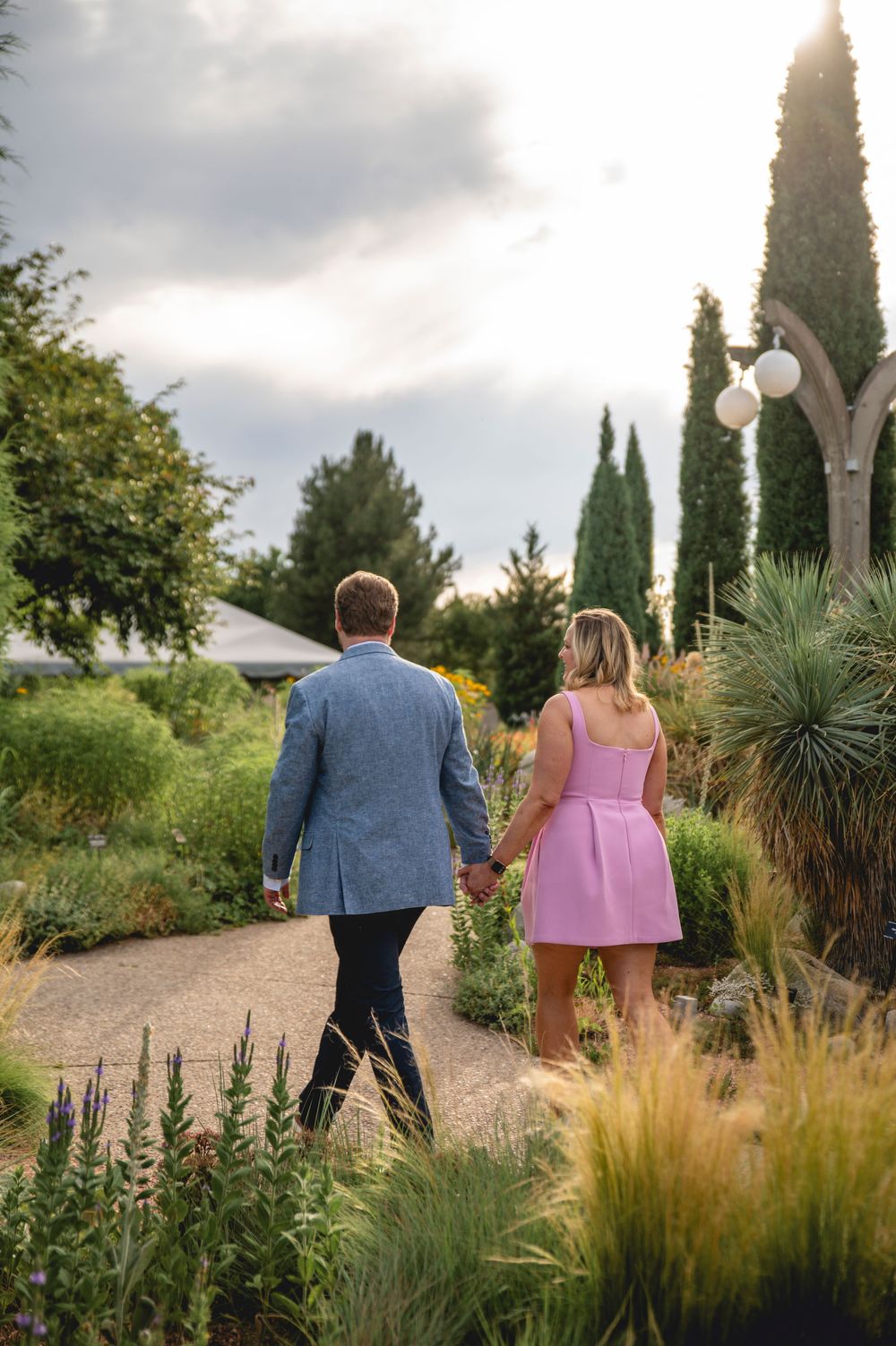 A couple walking together on a winding garden path surrounded by ornamental grasses and cypress trees at sunset.