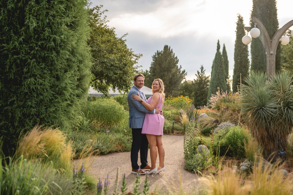A couple walks through a lush garden path surrounded by ornamental grasses and cypress trees on a sunny day.