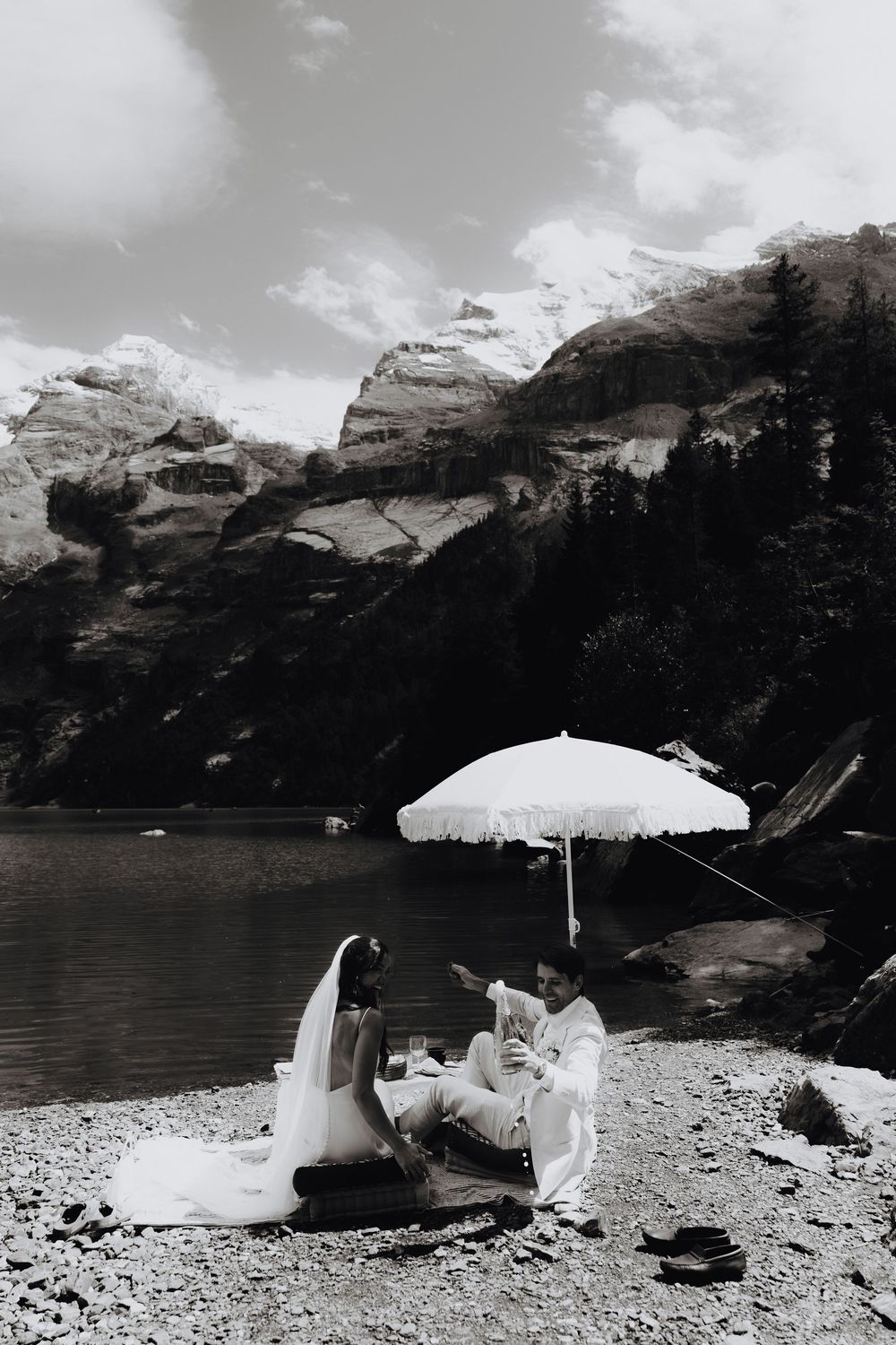 A beach umbrella shelters two people lounging by a serene mountain lake surrounded by rocky cliffs, captured in black and white.
