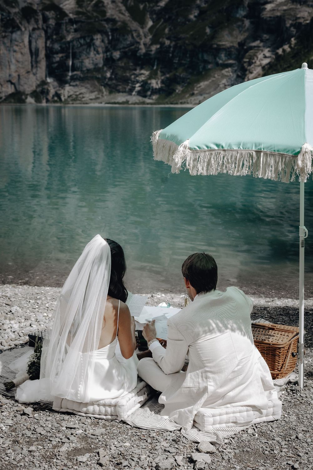 A couple in wedding attire sits by a turquoise lake under a mint green fringed umbrella during an intimate lakeside picnic.