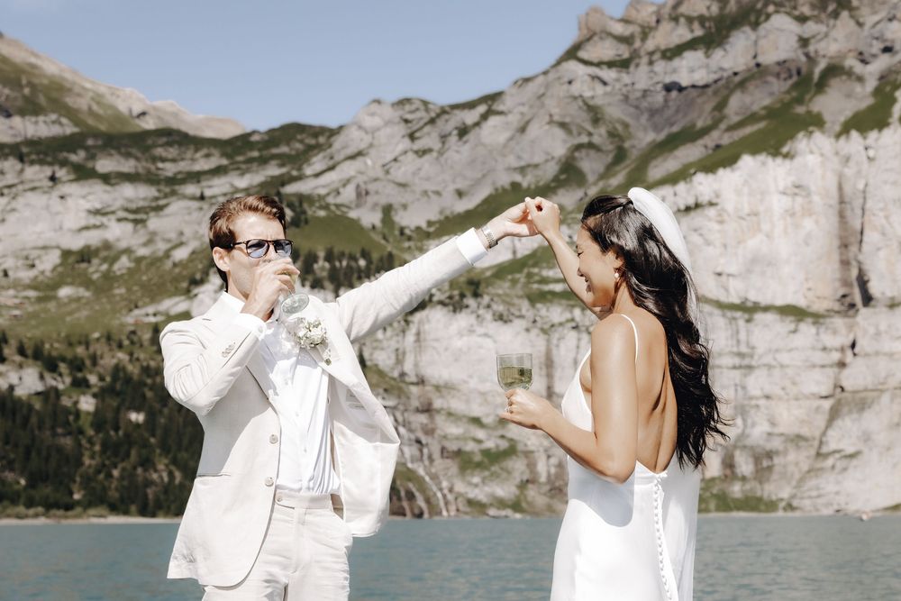A couple in white outfits poses together along a scenic mountain landscape with rocky cliffs in the background.