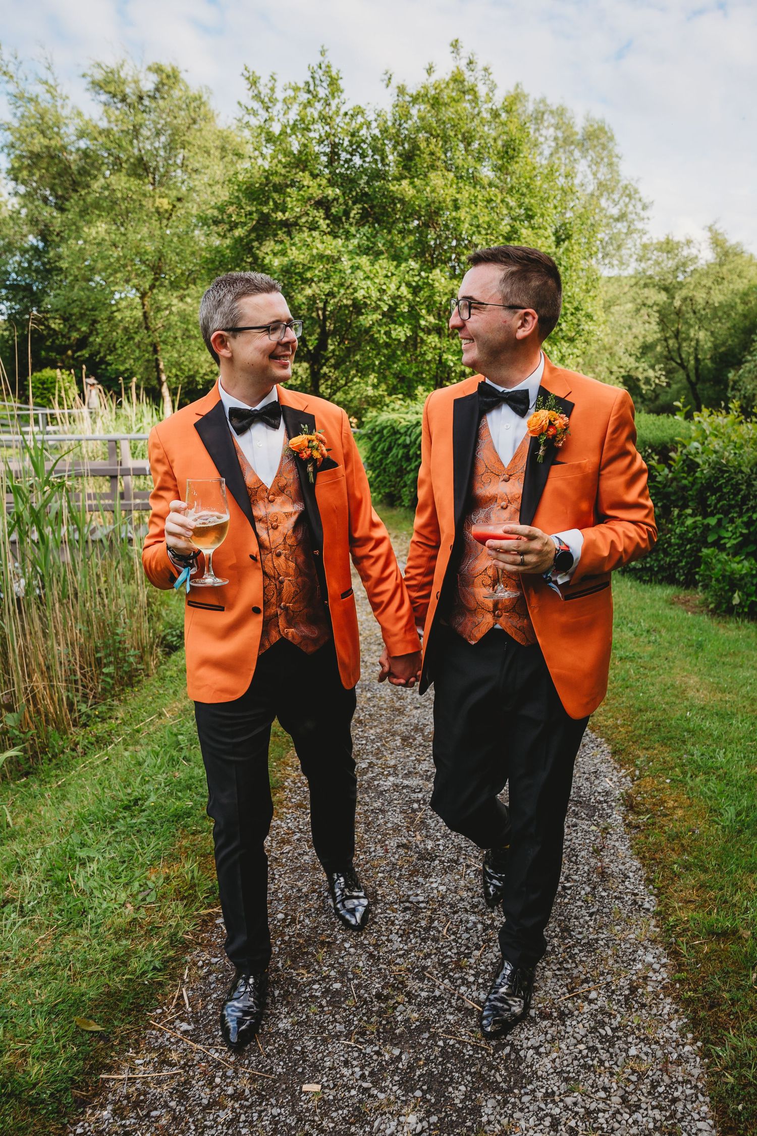 Two people in matching orange tuxedos and bowties walk together on a gravel path.