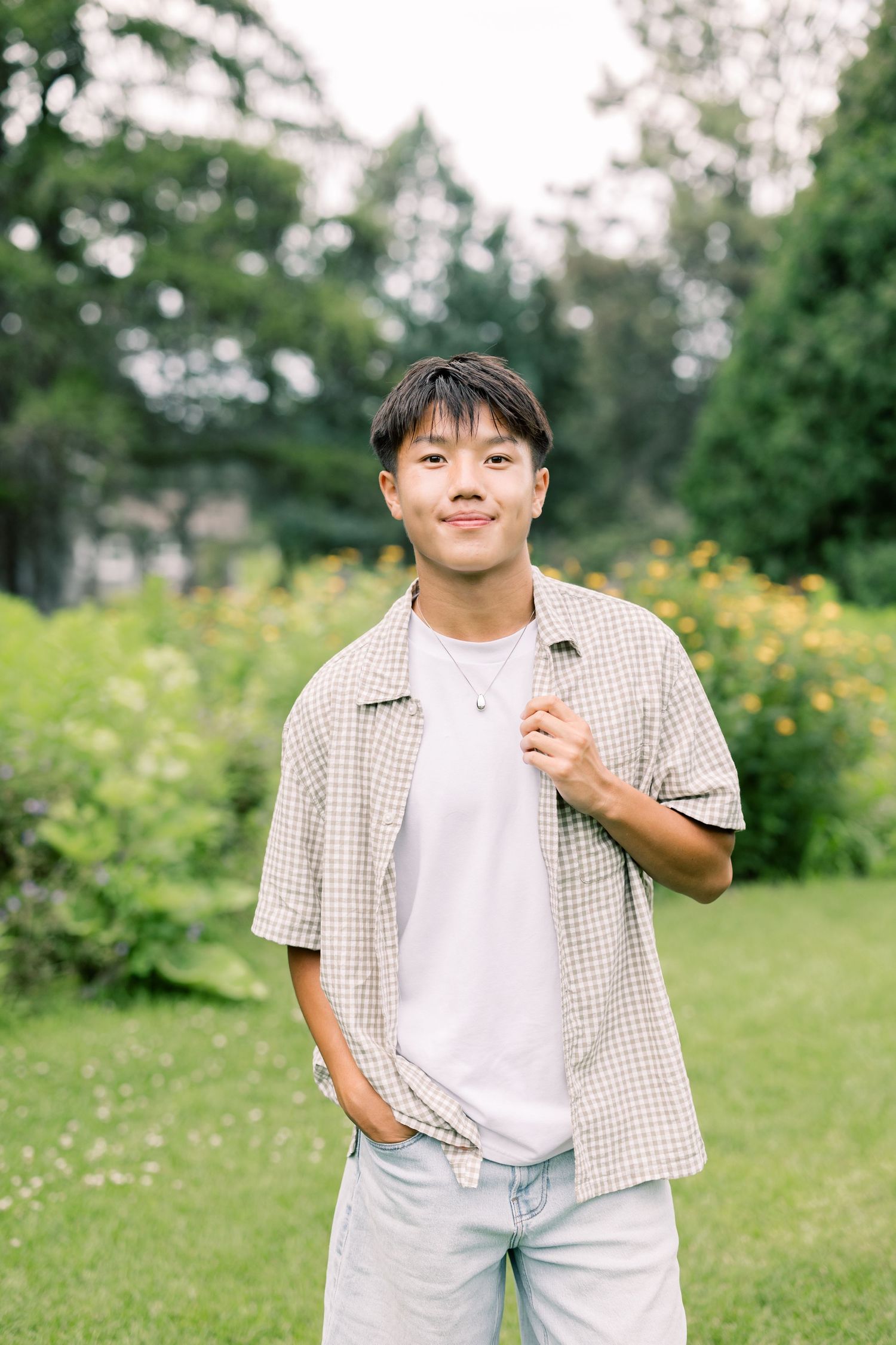 A young person wearing a white t-shirt and checkered shirt stands outdoors in a lush garden with yellow flowers.