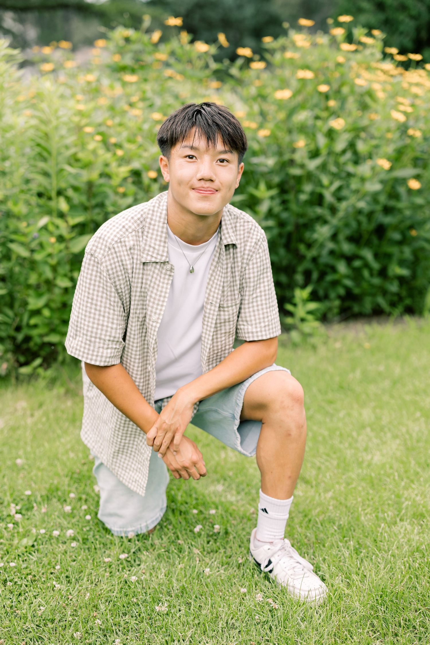 A child in a checkered shirt sits casually on grass surrounded by yellow flowers in the background.