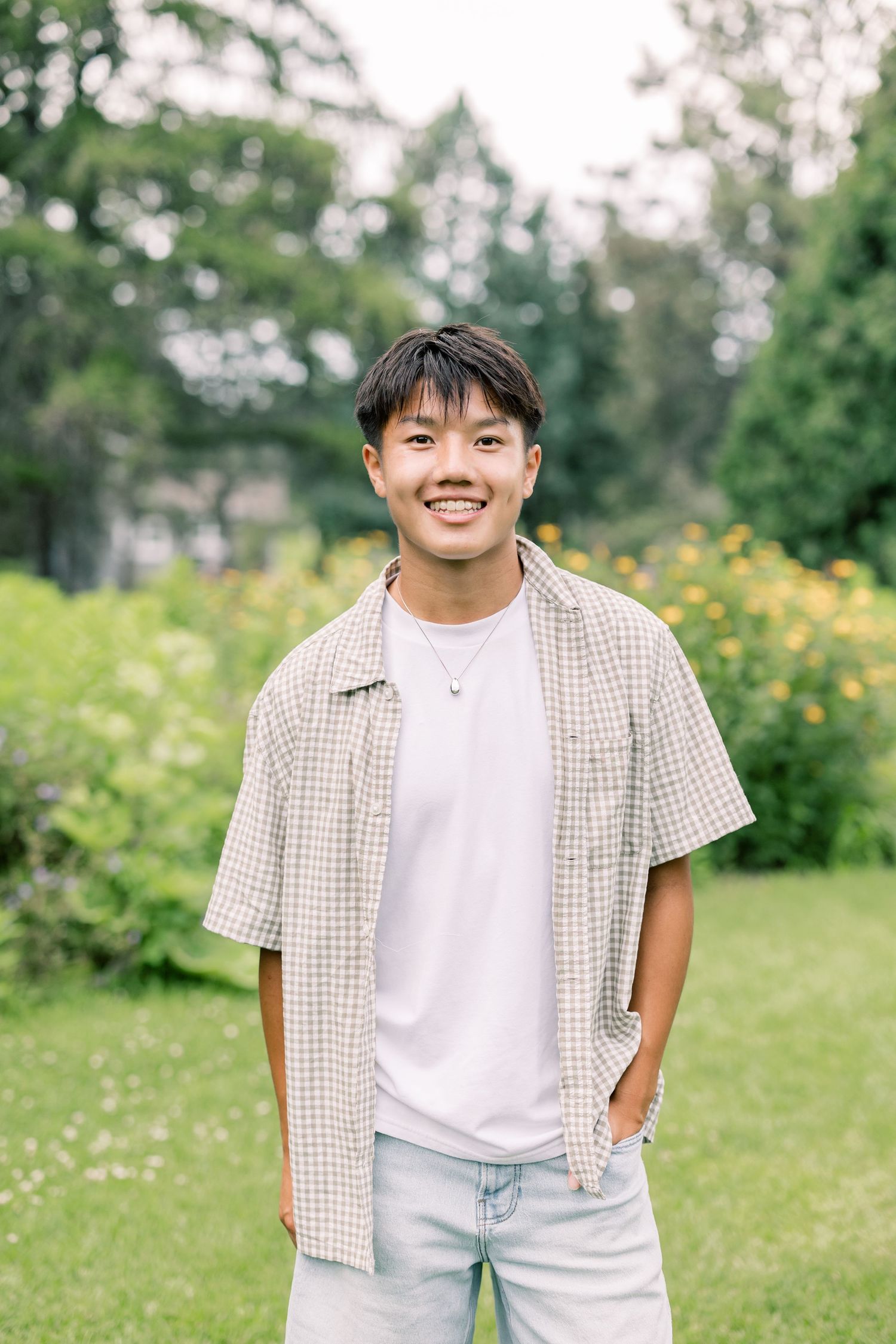 A person stands in a garden wearing a light checkered shirt and white t-shirt against a green nature backdrop.
