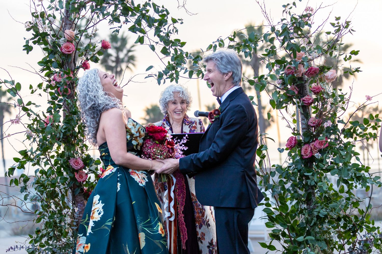 A couple laughs during an outdoor wedding ceremony surrounded by pink floral arrangements and greenery.