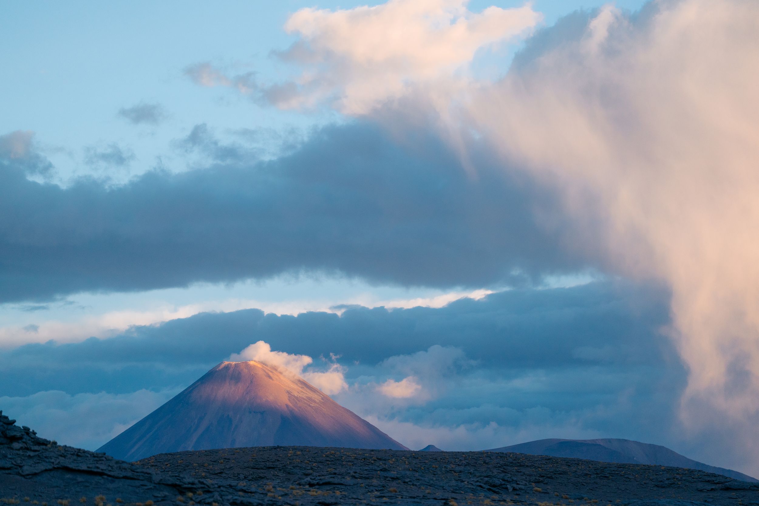 Mountain peak glowing pink in sunset light with dramatic clouds and blue sky over rugged landscape.