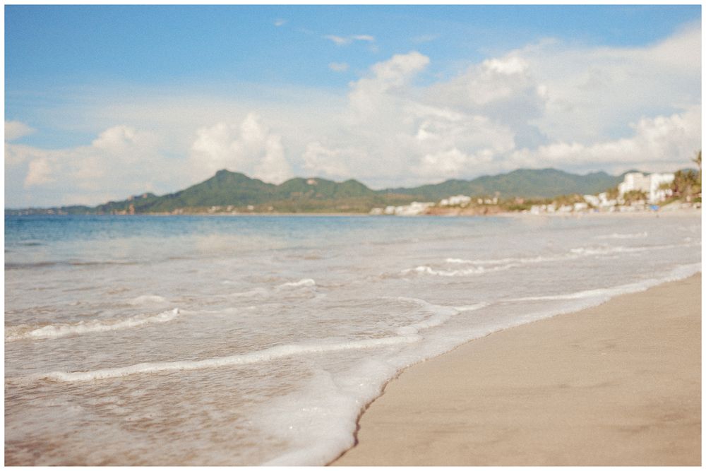 Waves gently roll onto a sandy beach as mountains rise in the distance under a cloudy sky.