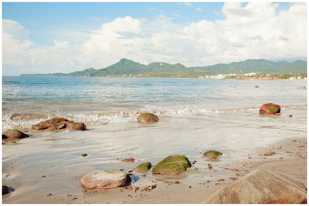 Waves gently roll onto a sandy tropical beach with distant mountains and scattered rocks along the shoreline.