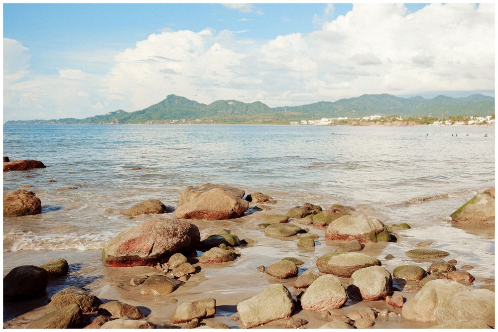 Large boulders scattered along tropical beach with turquoise water and distant mountains.