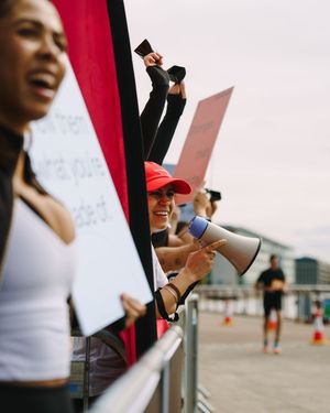 A person in a white top holding a megaphone and protest sign stands against a bright red background.