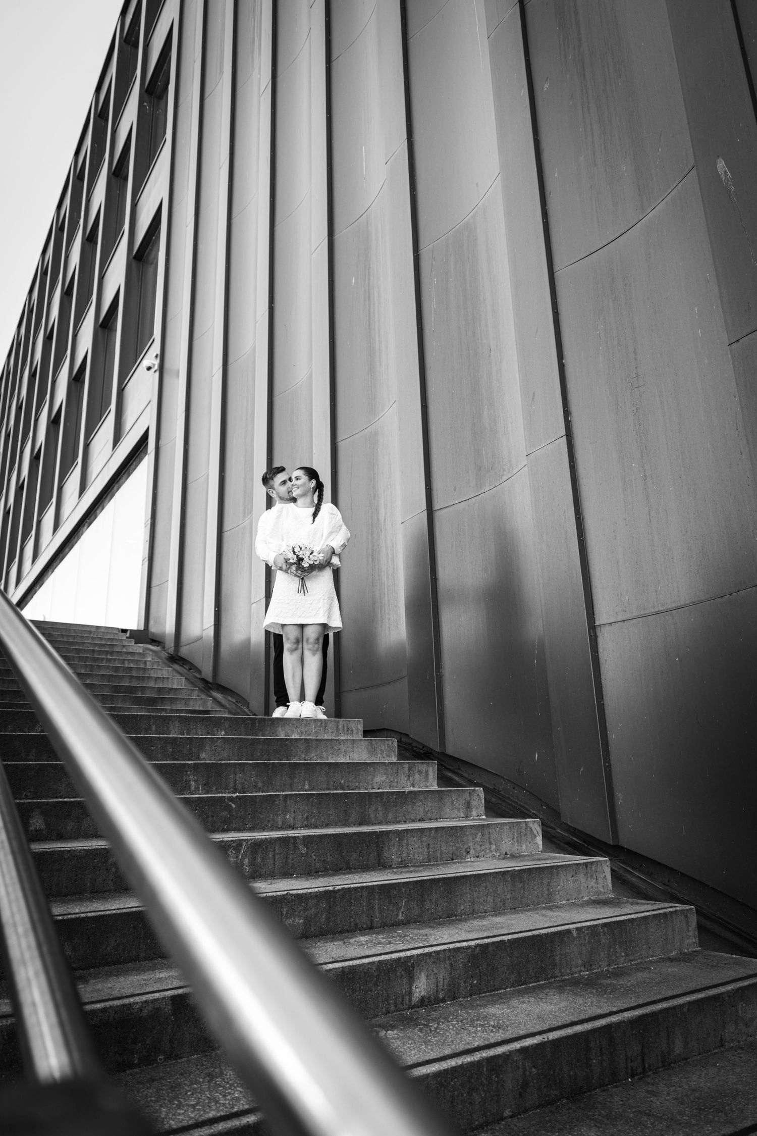 Black and white photo of wedding couple embracing on outdoor stairs beside a modern building wall.
