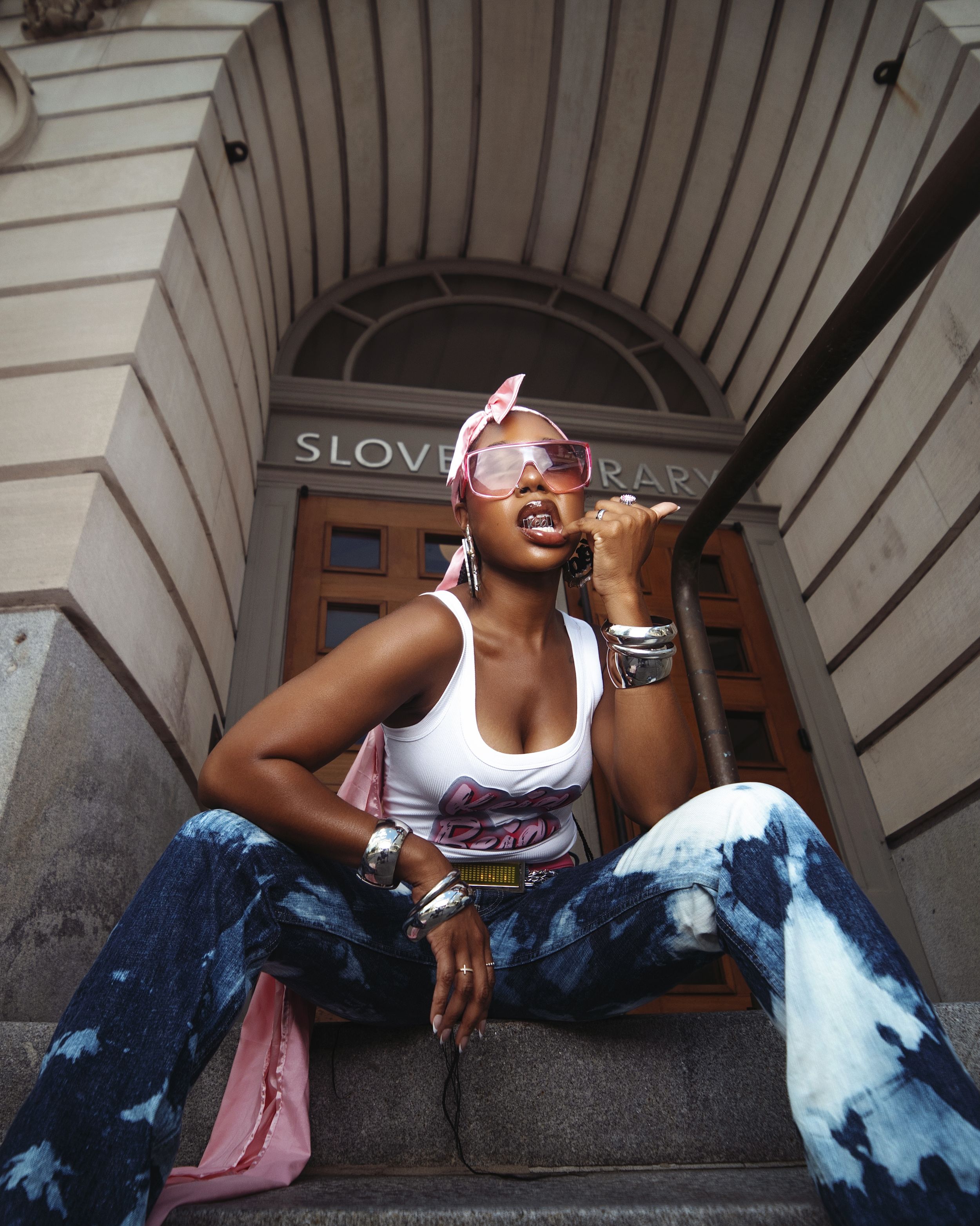 A person in bleached denim and white tank top sits casually on stone steps in front of an ornate building entrance.