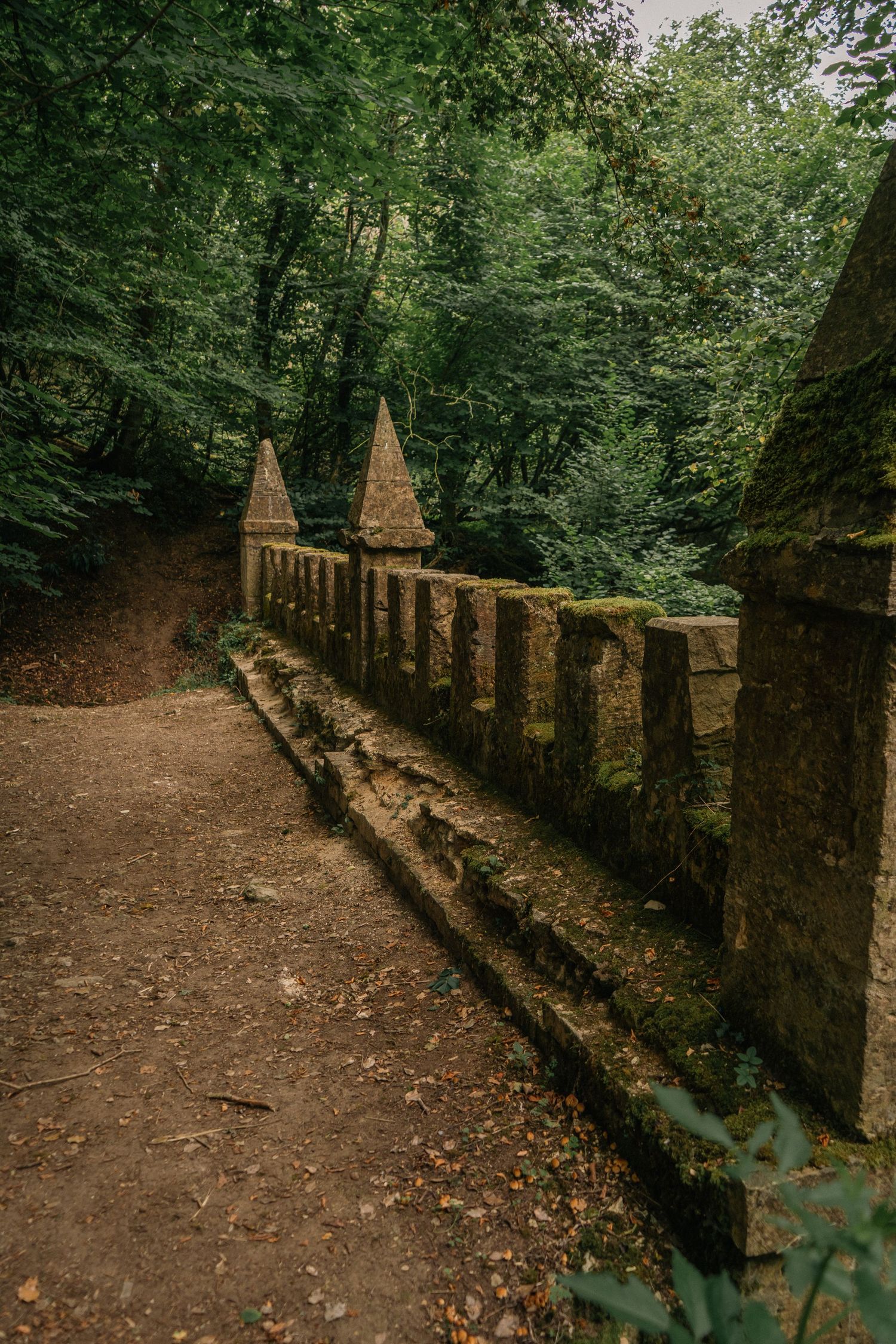 Author novel promotional photoshoot in Cotswold. Ancient stone bridge with decorative spires surrounded by lush green forest on a dirt path.