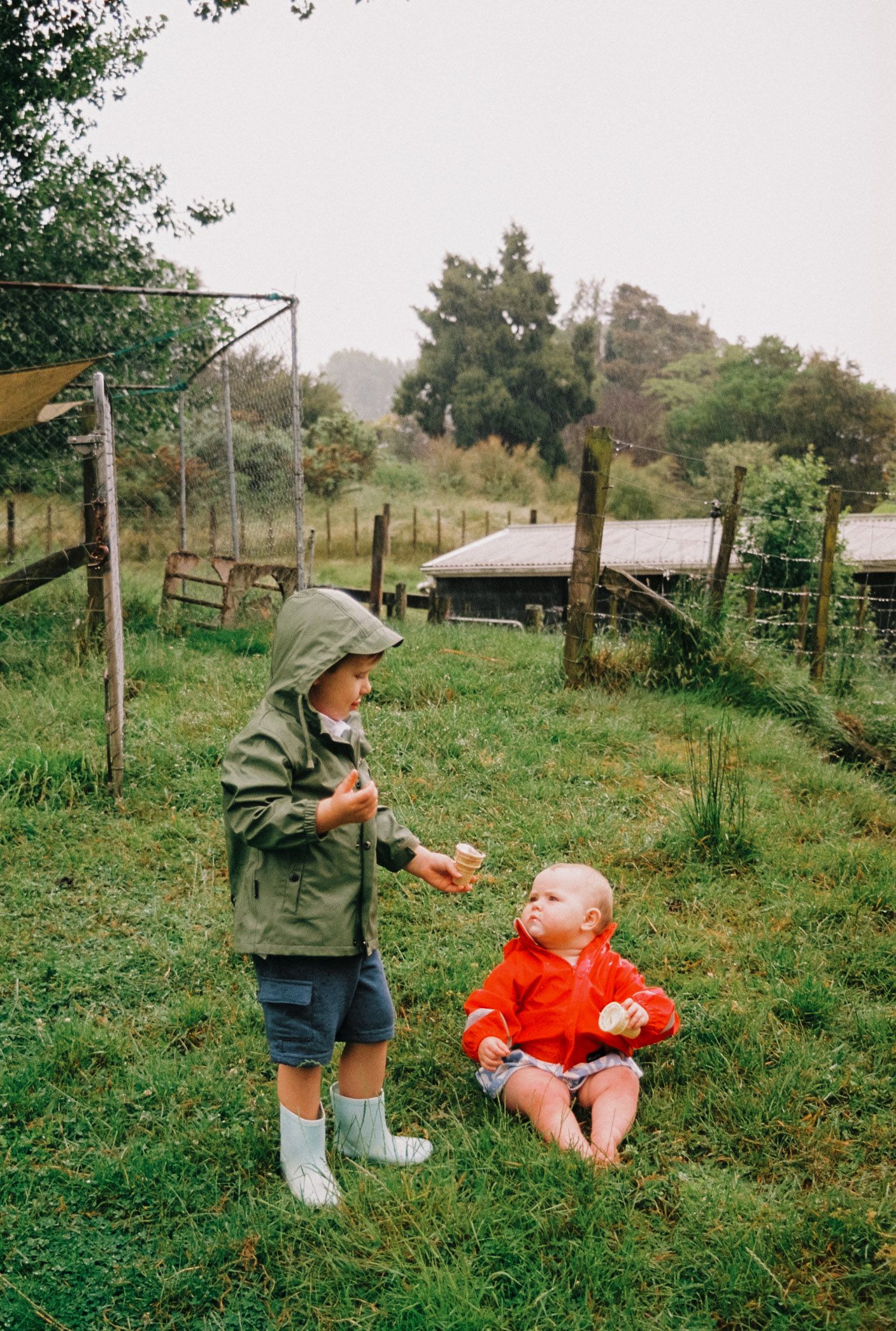 Two children in rain gear play together on grassy farmland with wooden structures and trees in the background.