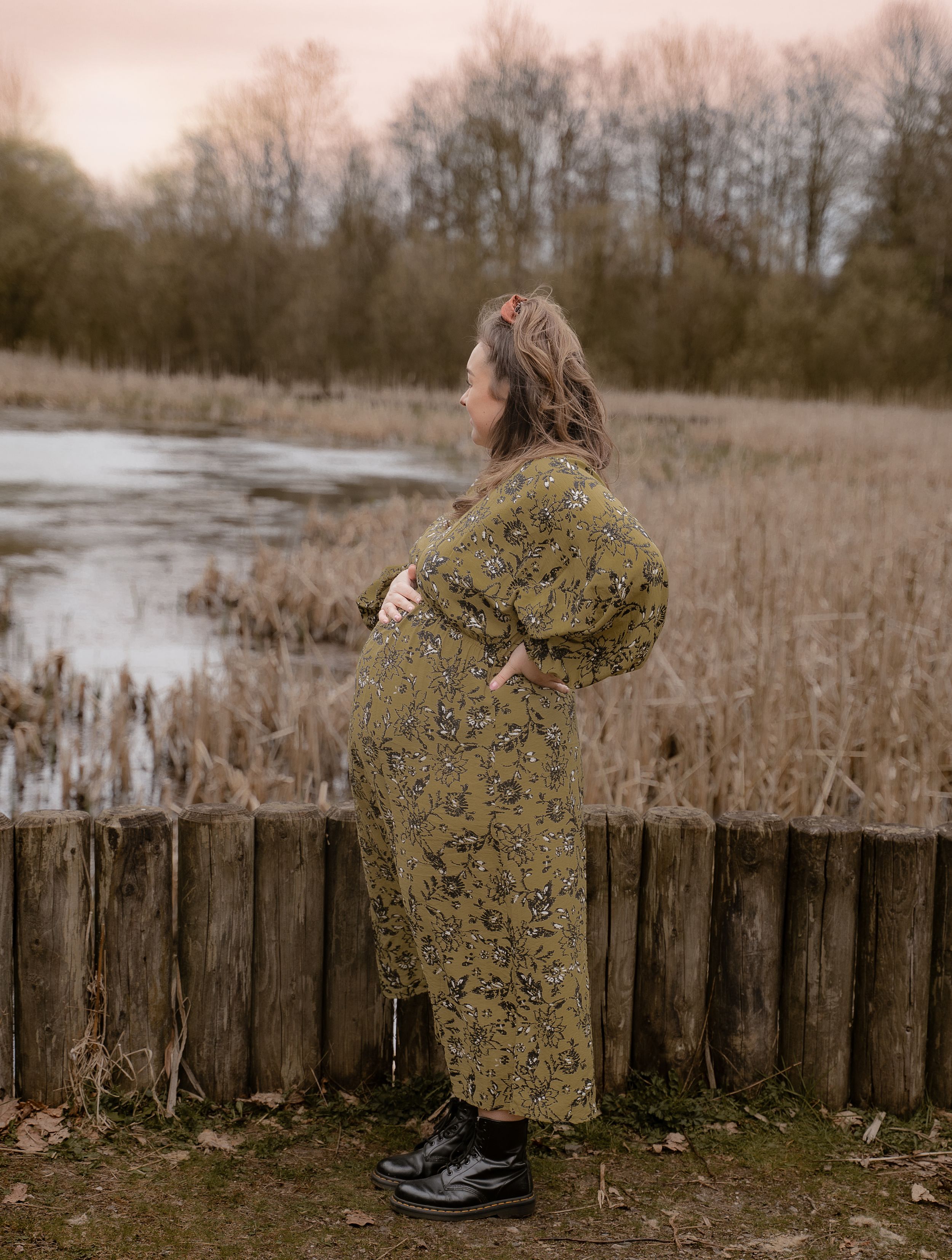 A pregnant woman in a floral dress stands at a wooden fence overlooking a serene lake surrounded by bare winter trees.