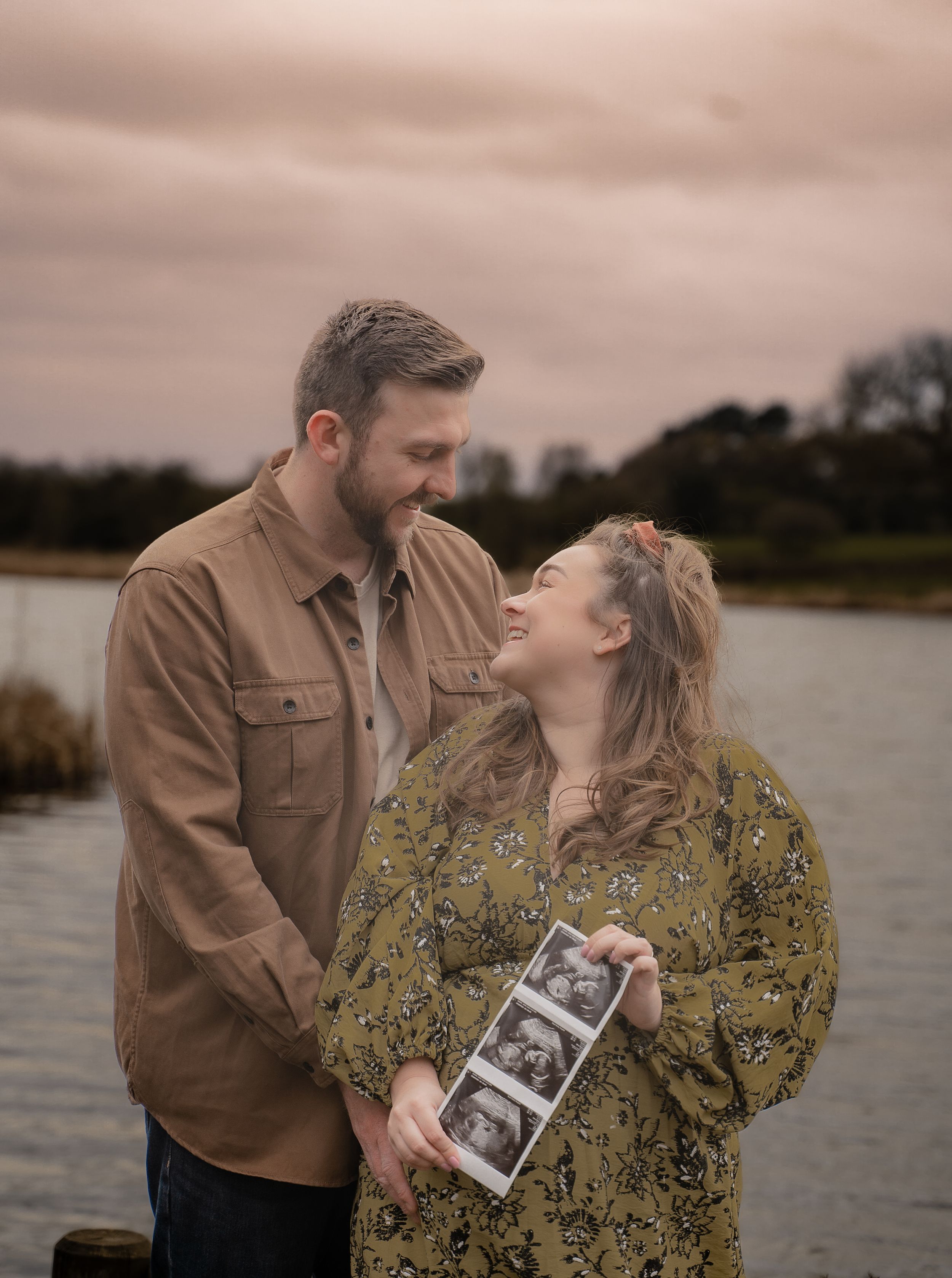 A couple shares a joyful moment by a lake while holding an ultrasound photo during a sunset pregnancy announcement shoot.
