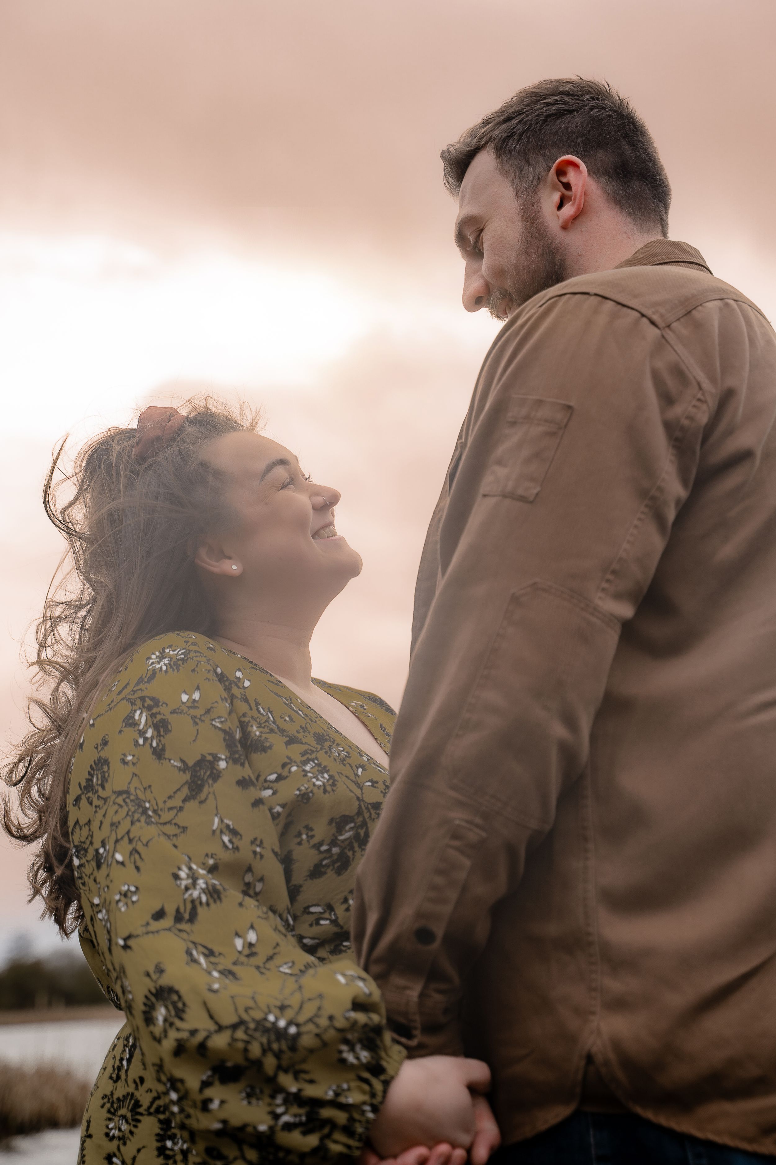 A couple shares an intimate moment outdoors, with her in a floral dress and him in a brown jacket at sunset.