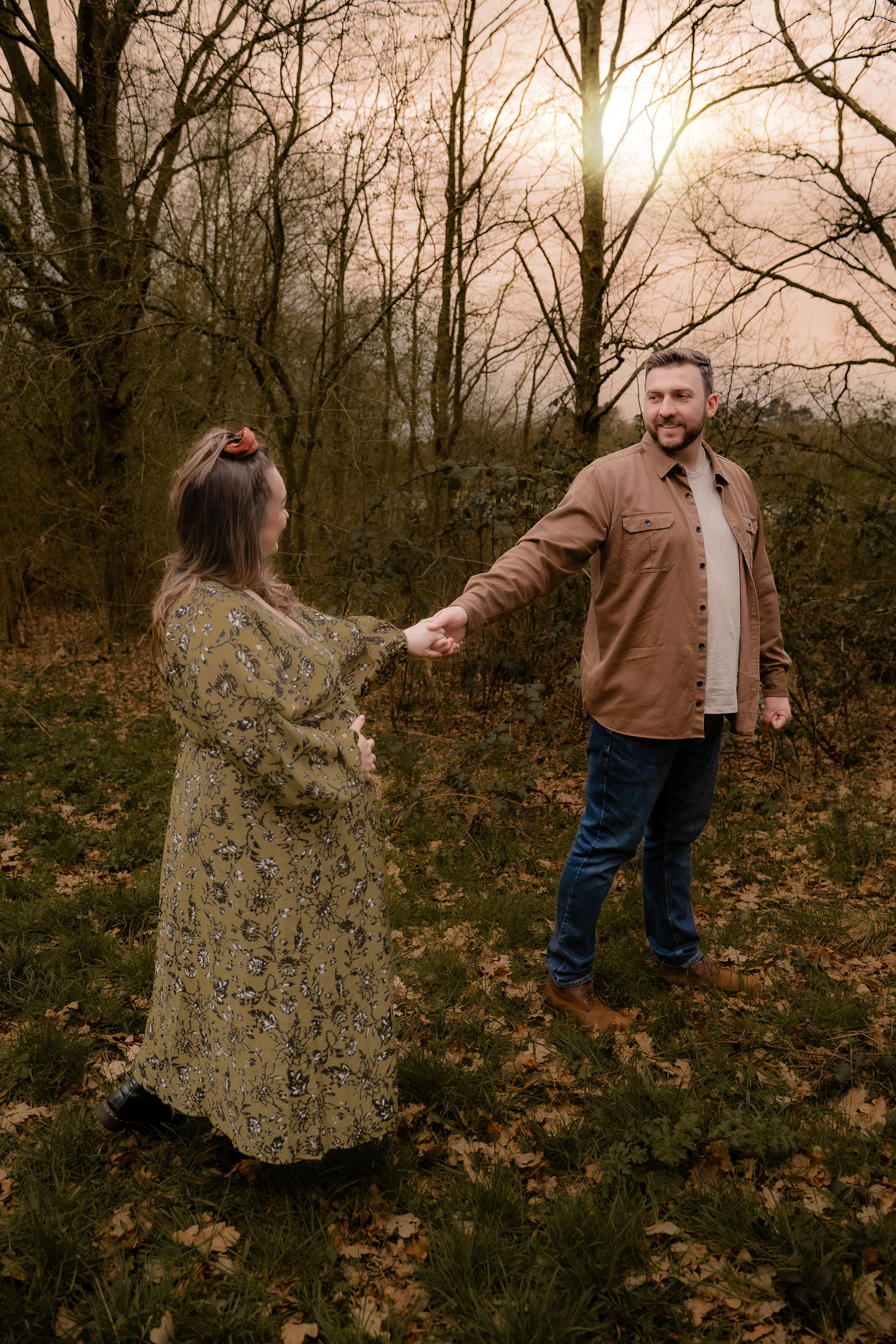 A couple dances together in a wooded area during autumn with fallen leaves scattered on the ground at sunset.
