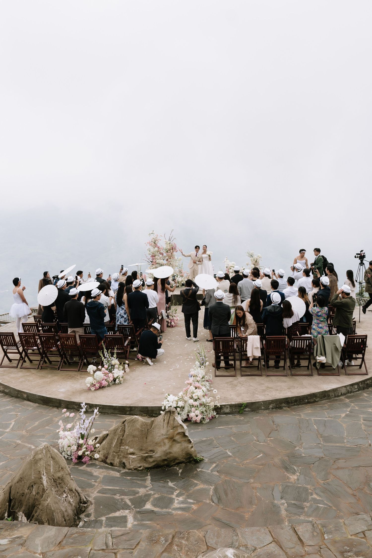Aerial view of an outdoor wedding ceremony on a stone terrace with guests holding parasols and floral aisle decorations.