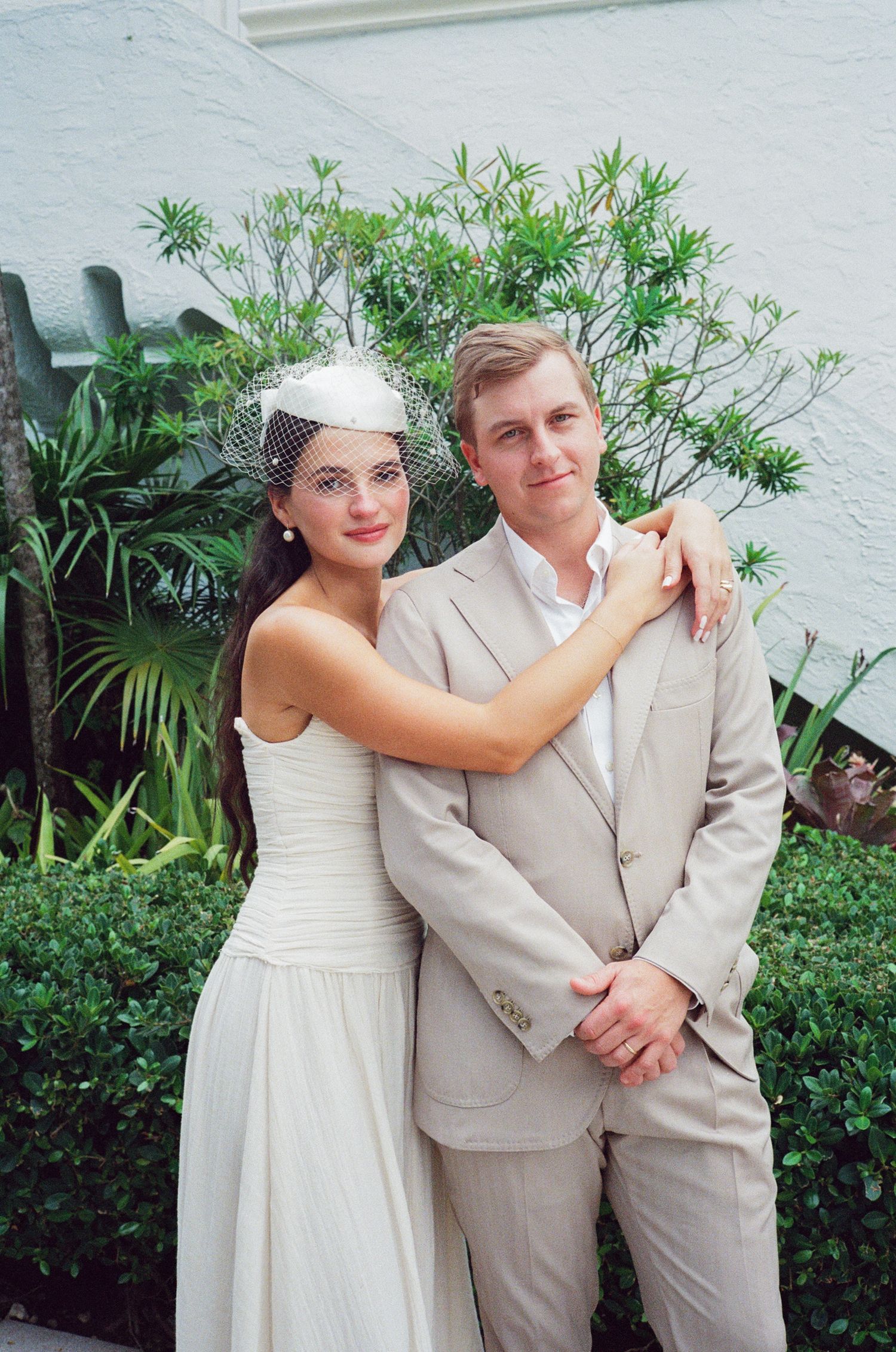 A bride in white dress and groom in beige suit pose playfully together against a garden backdrop with tropical plants.
