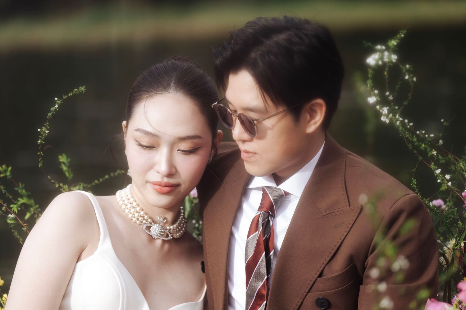 Close-up of elegant bride and groom in brown suit surrounded by lush greenery and colorful flowers outdoors.