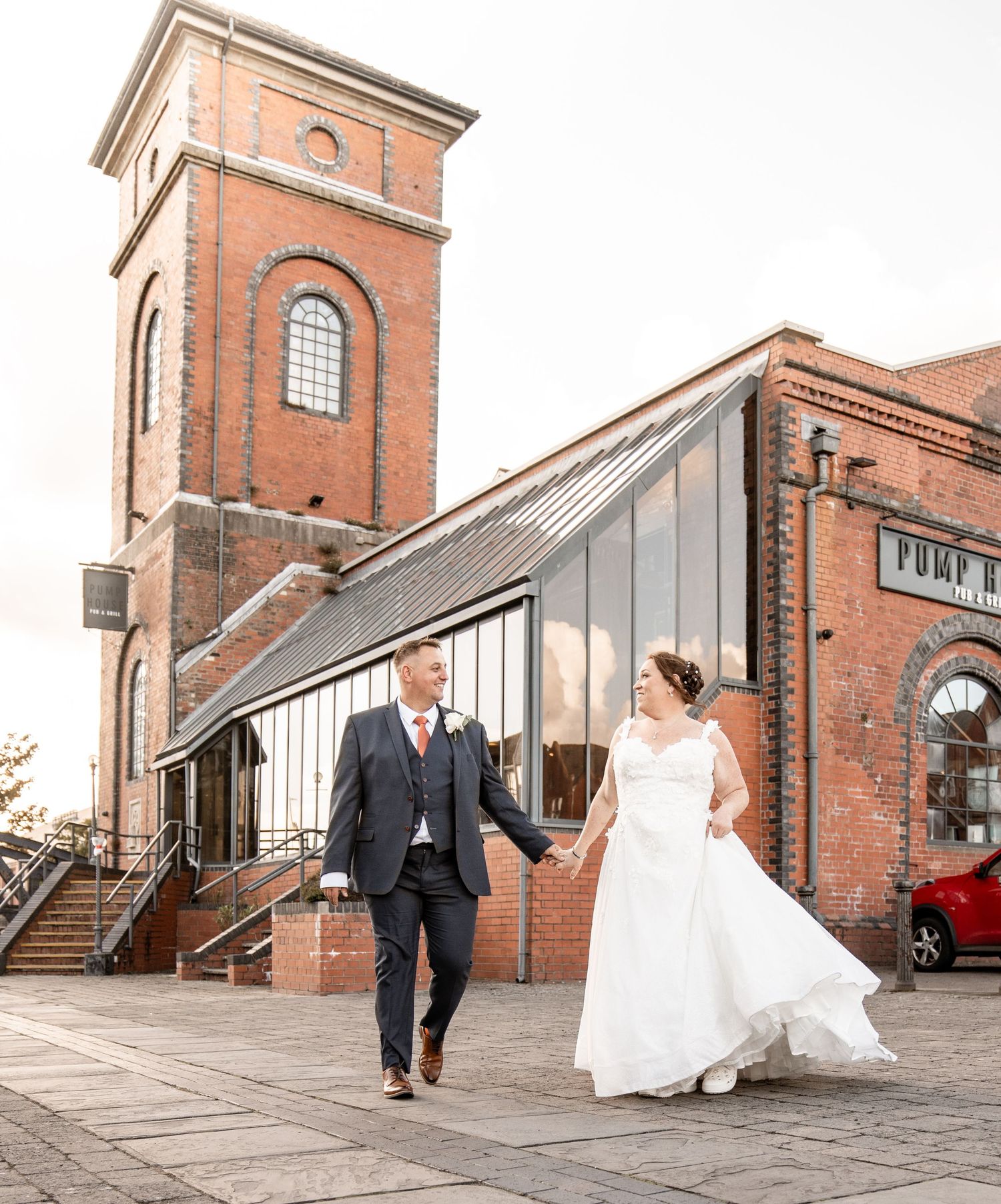 A wedding couple shares joyful moments outside a historic brick industrial building with tall windows and stone steps.