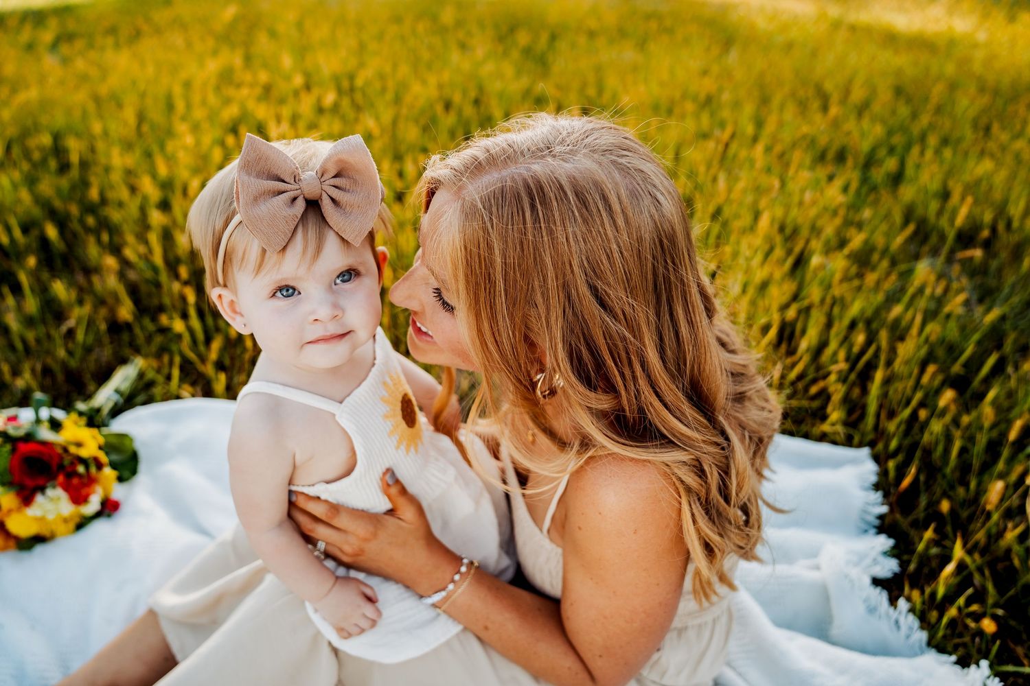 A parent shares an intimate moment with their child while lying in a sunny meadow with white floral decor around them.