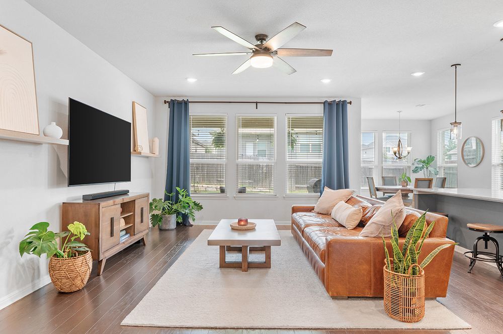 Modern living room with brown leather sectional, ceiling fan, wall-mounted TV, and blue curtains against white walls.