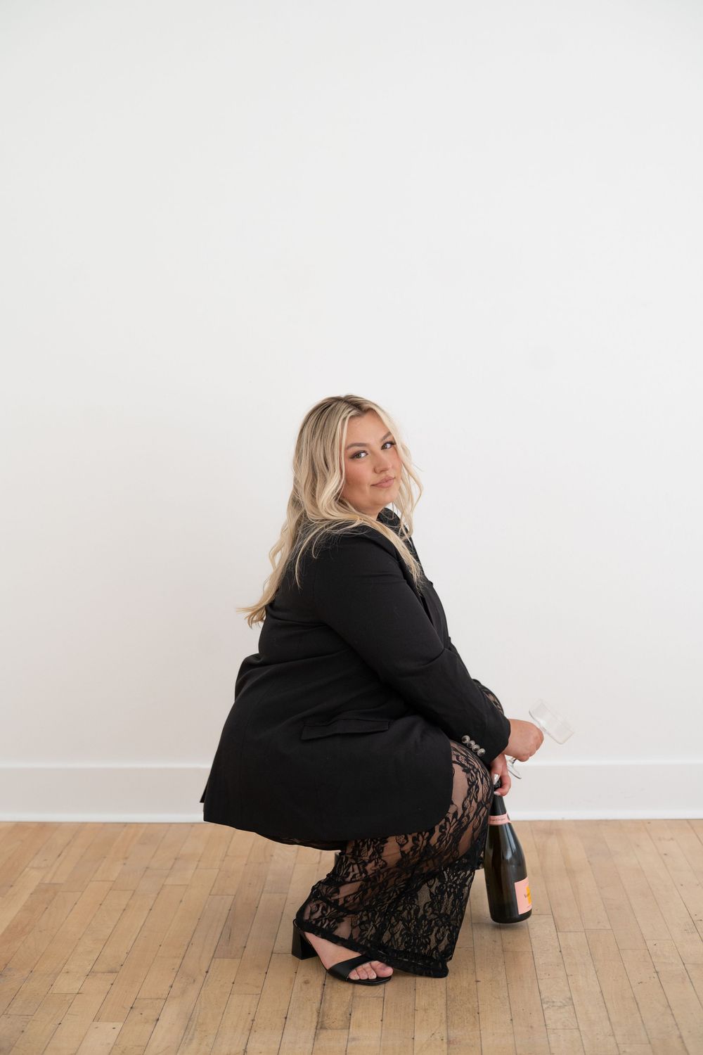 Figure in black attire crouching against white wall with wooden flooring.