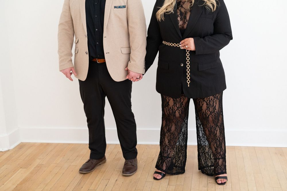 A stylish pair in contrasting outfits standing against a white wall.