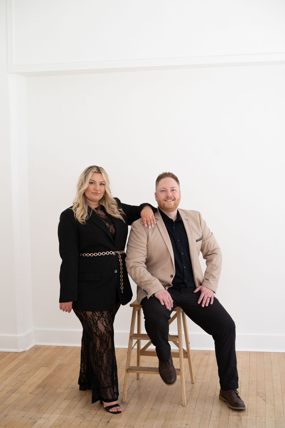 A couple posing in a bright studio space with one seated on a wooden stool.