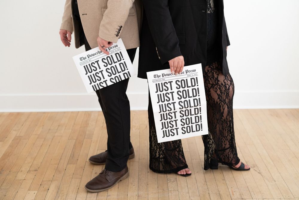 Two people holding 'JUST SOLD' signs while standing on hardwood flooring.