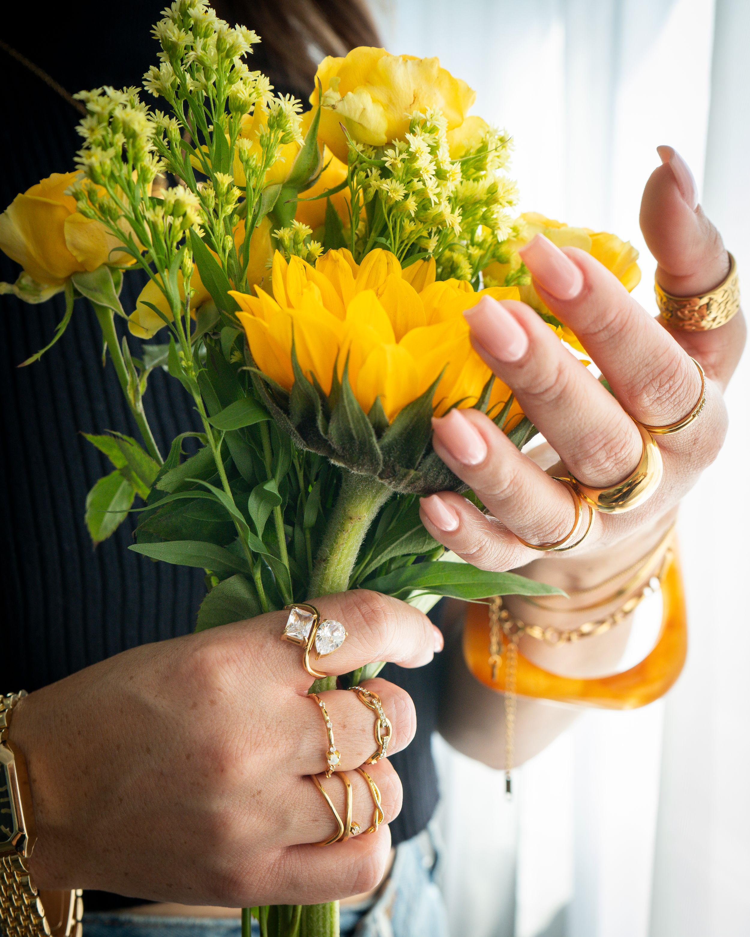Hands holding yellow flowers, adorned with multiple gold rings, bracelet, and orange bangle.