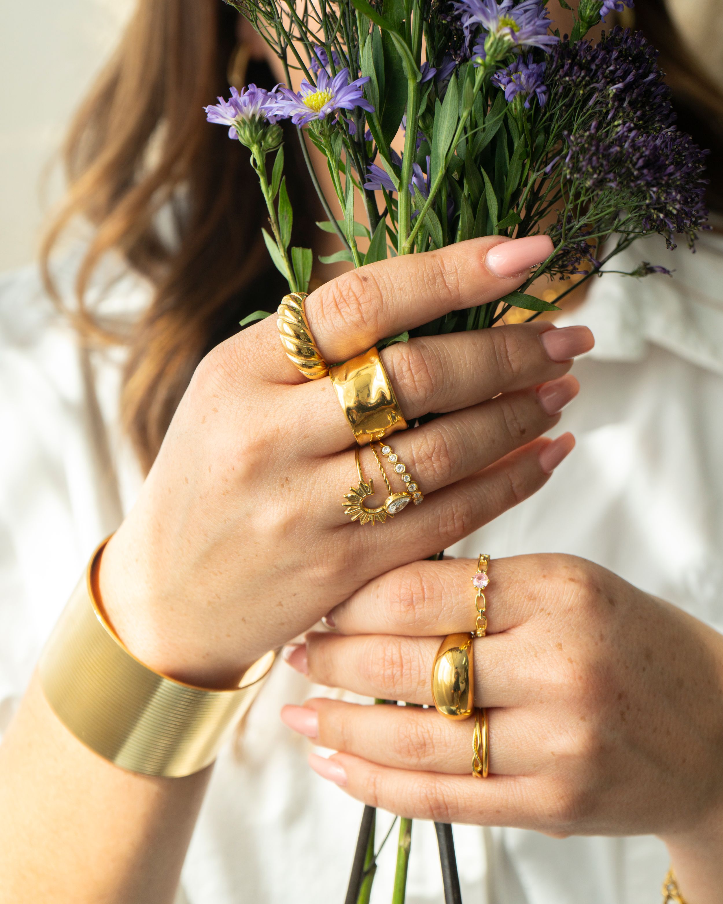 Close-up of hands holding purple flowers, stacked with gold rings and a wide cuff bracelet.