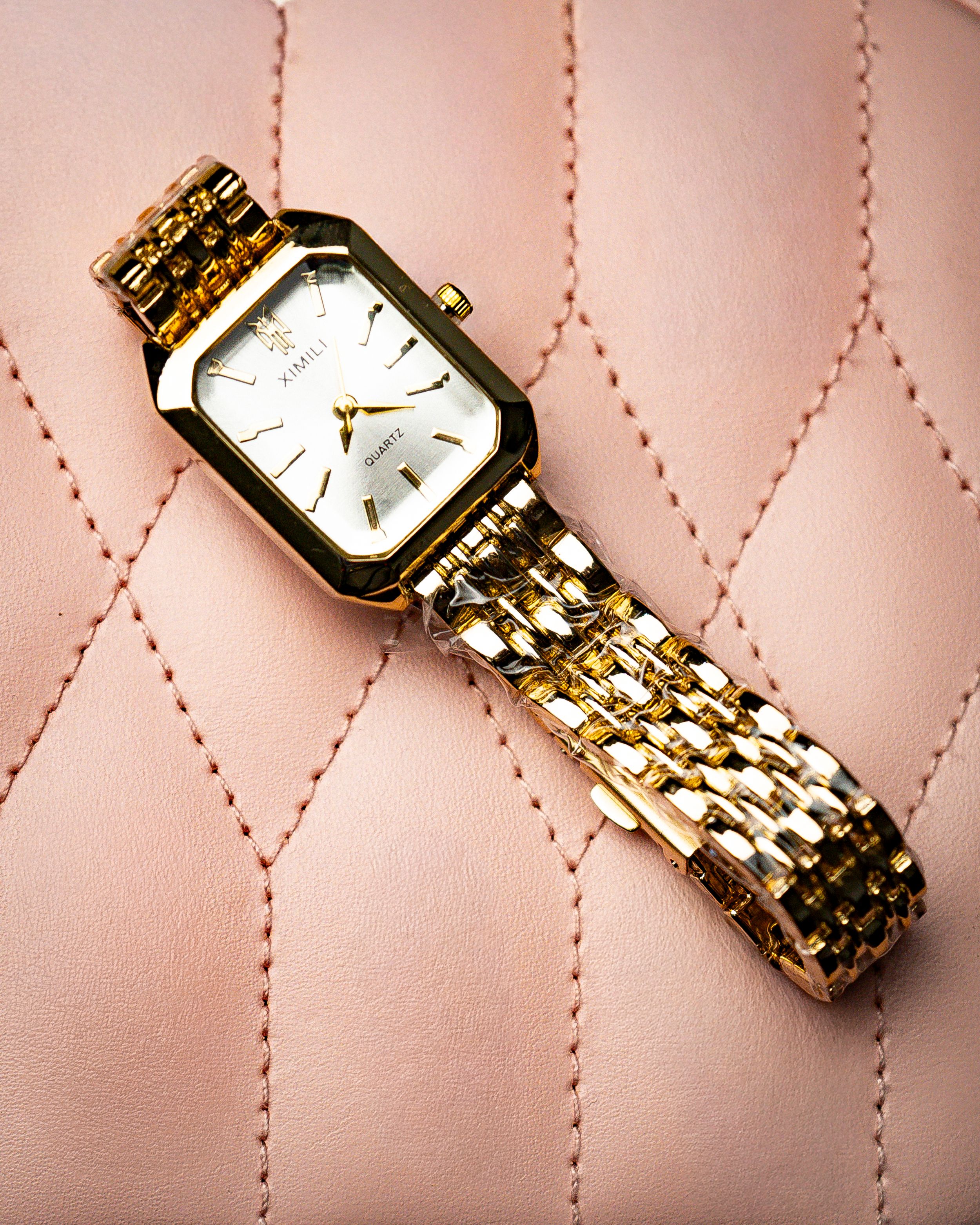 Gold wristwatch with a rectangular face on a quilted pink surface.