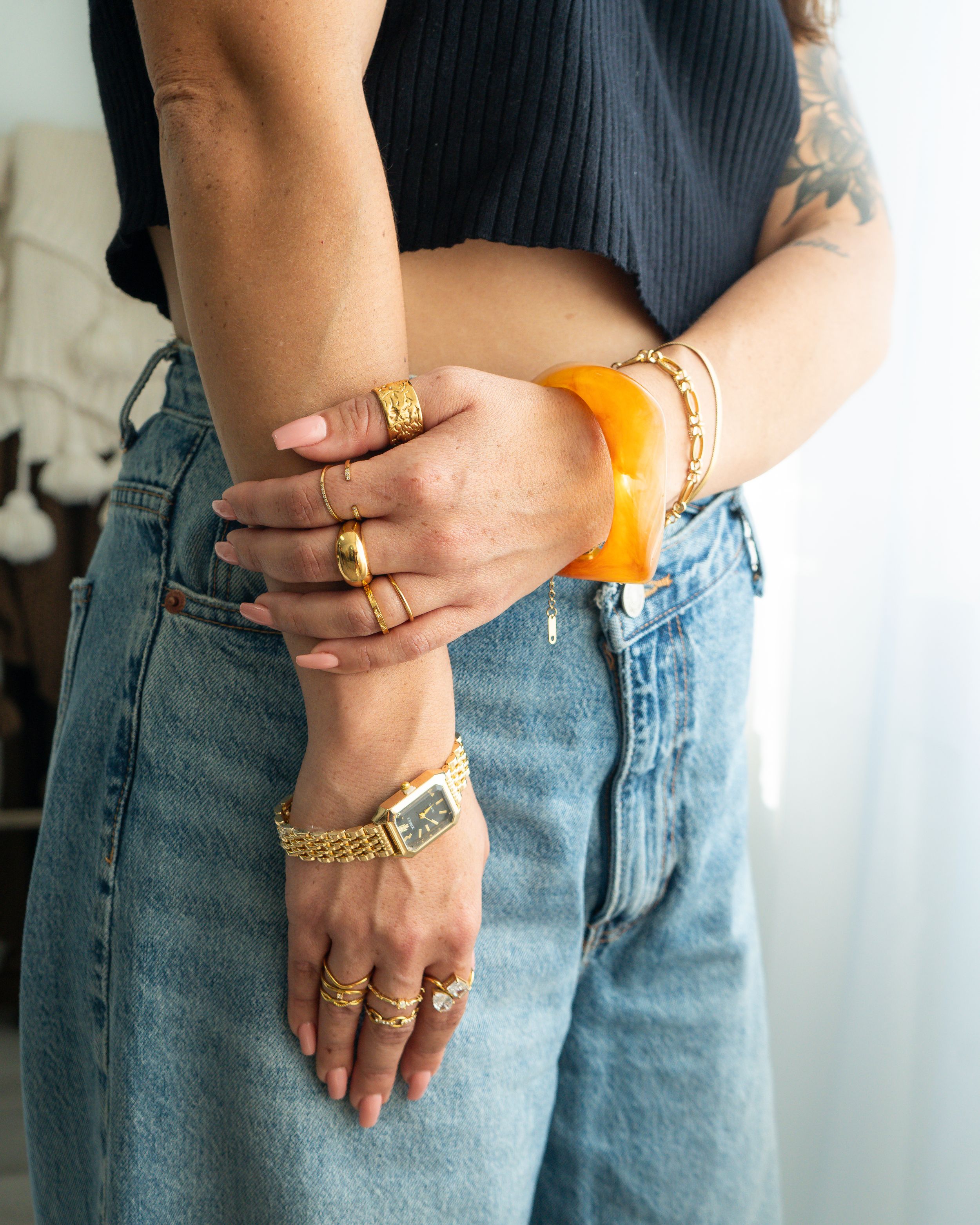Person in jeans and black top crossing arms, showing gold rings, watch, and chunky orange bracelet.