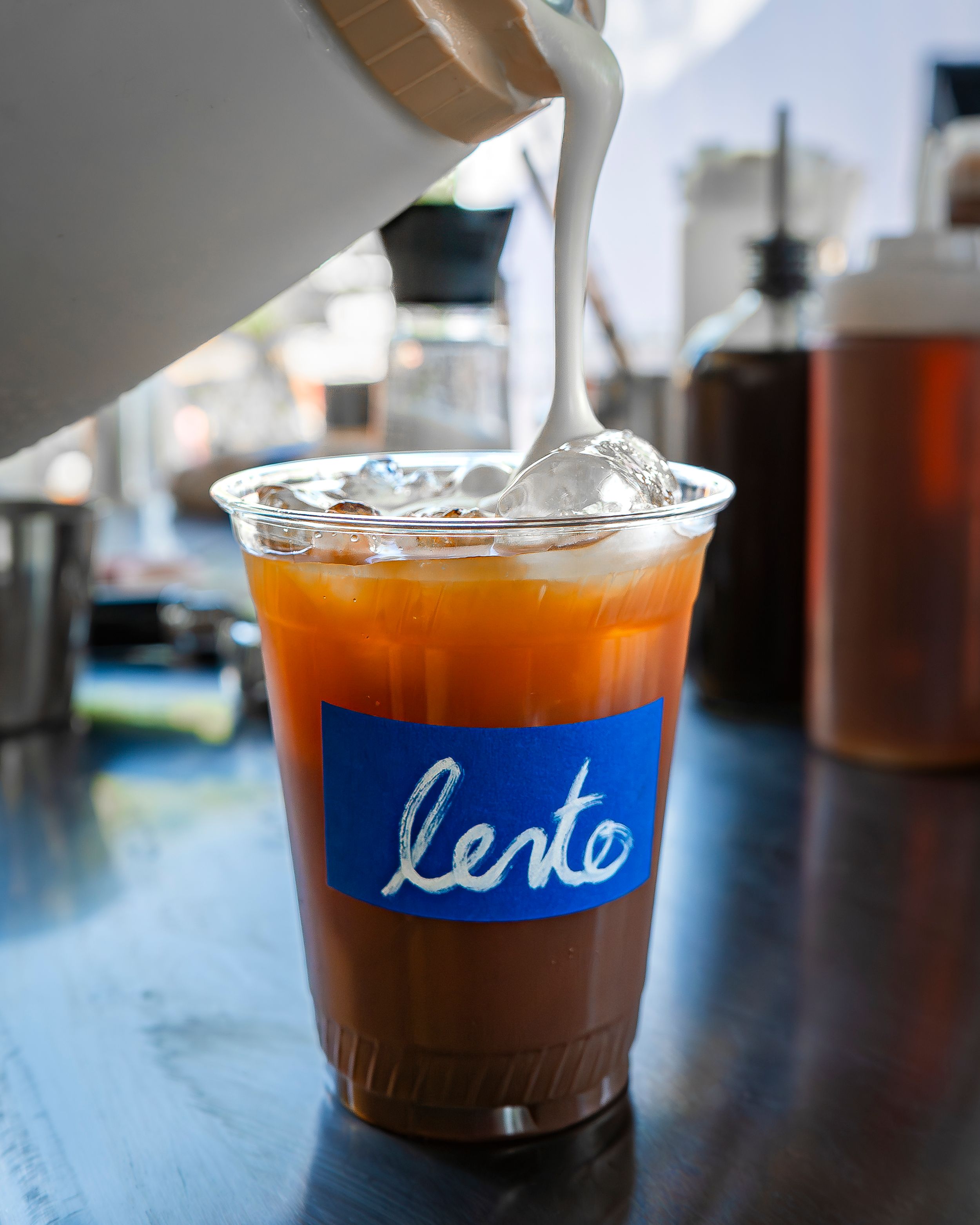 Milk being poured into an iced coffee in a “lento” cup on a cafe counter.