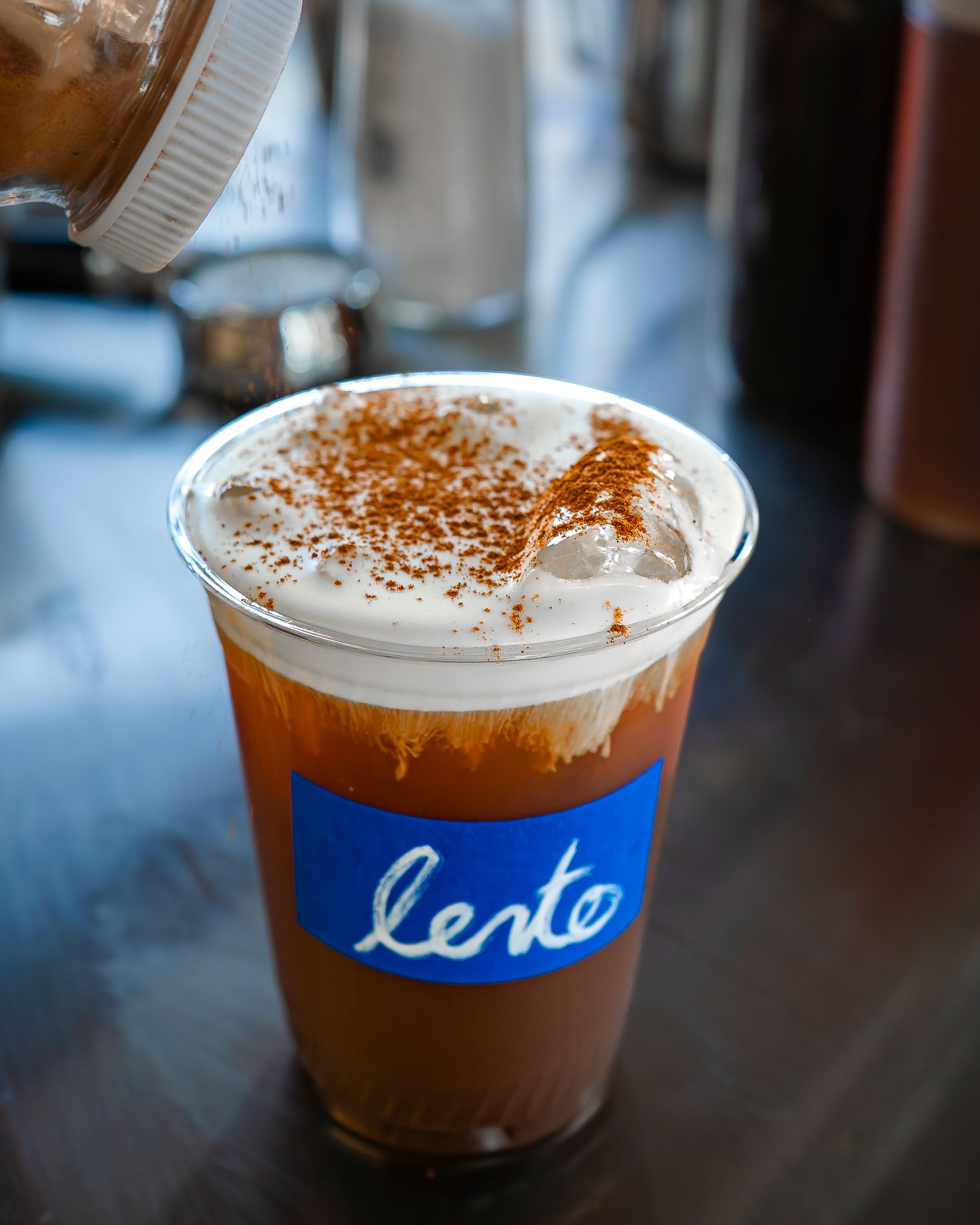 Iced coffee in a “lento” cup topped with foam and sprinkled spice on a cafe counter.