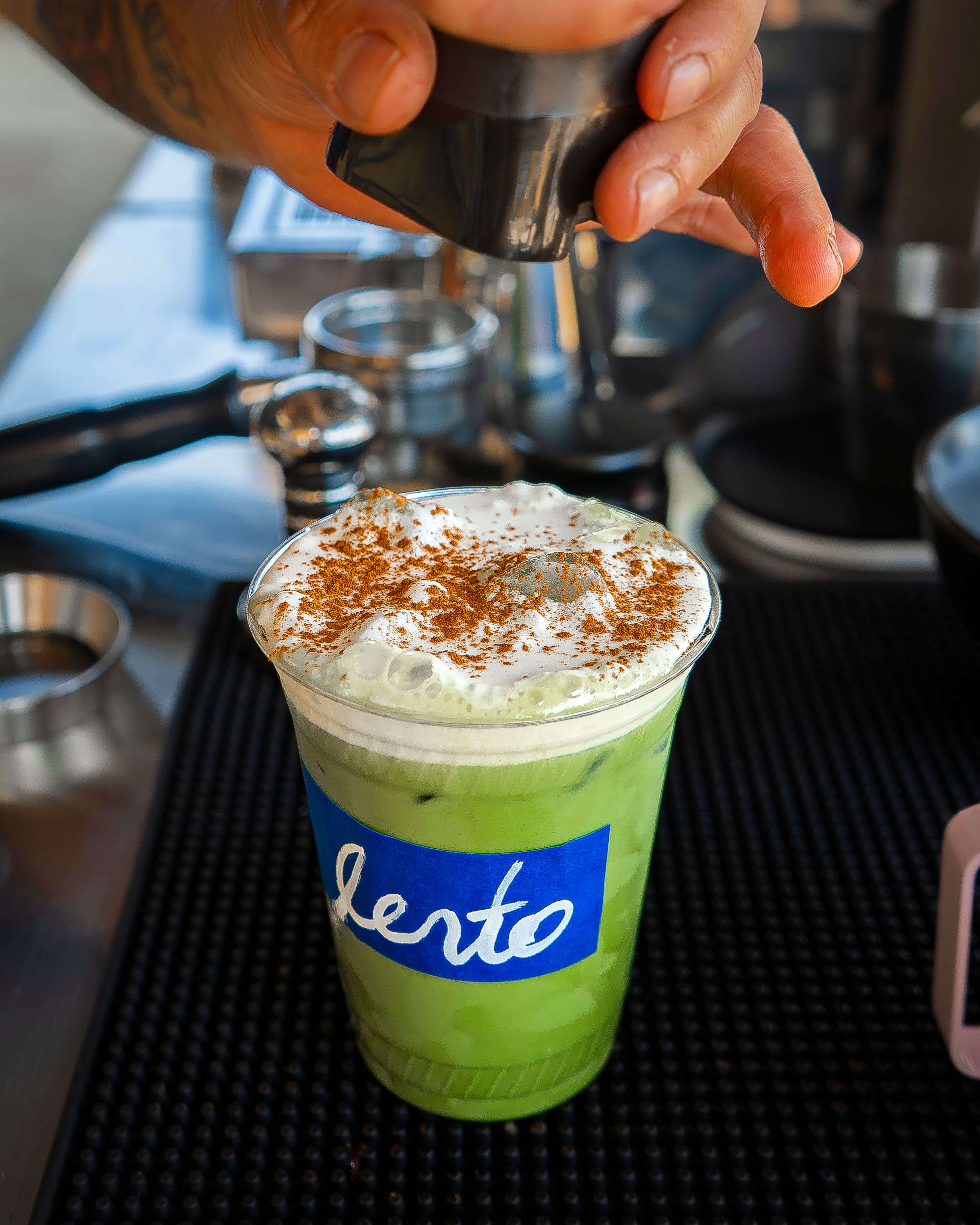 Barista sprinkles spice over whipped-topped iced matcha drink in a “lento” cup.
