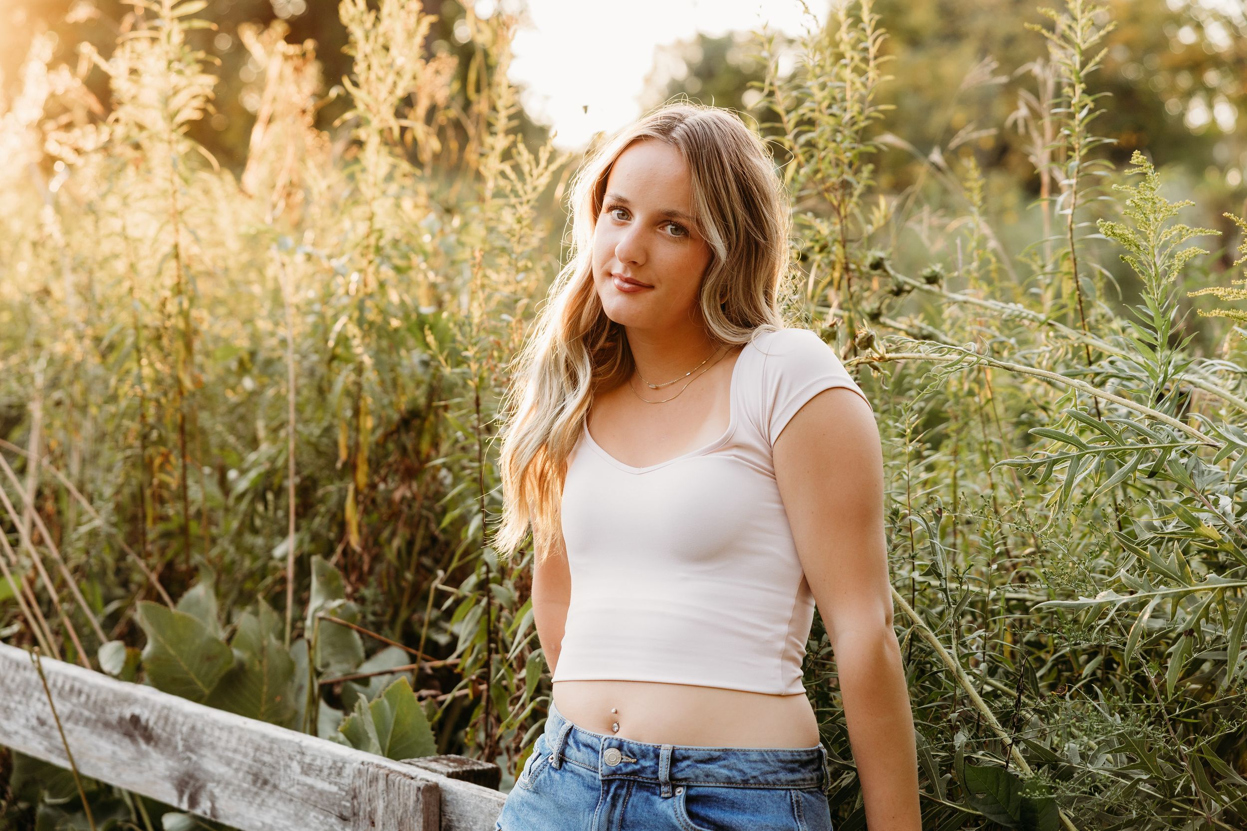 Someone in a white crop top and jeans poses outdoors among tall grasses during golden hour lighting.