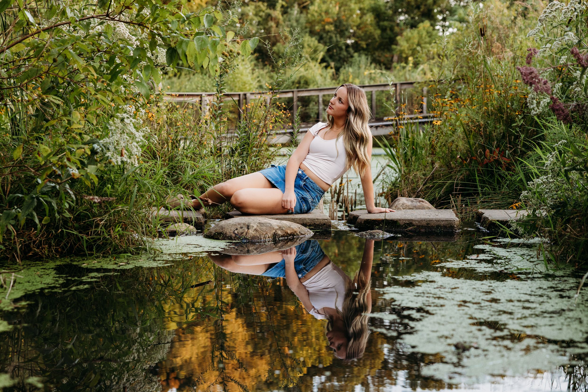 People enjoying a summer day along a serene pond with lush green foliage and a rustic wooden bridge in the background.