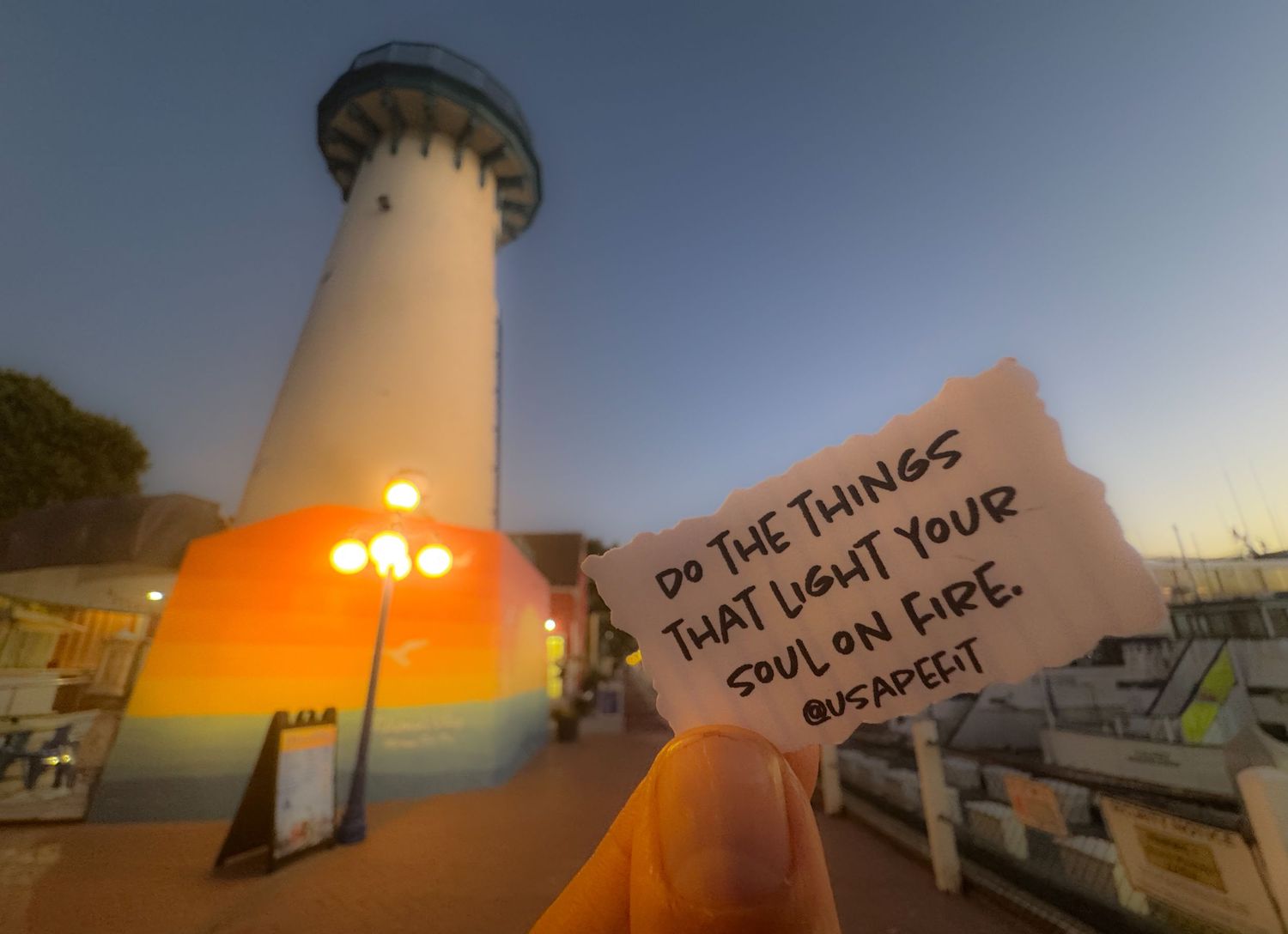Lighthouse illuminated in warm orange glow against a vibrant sunset sky with handwritten message visible.