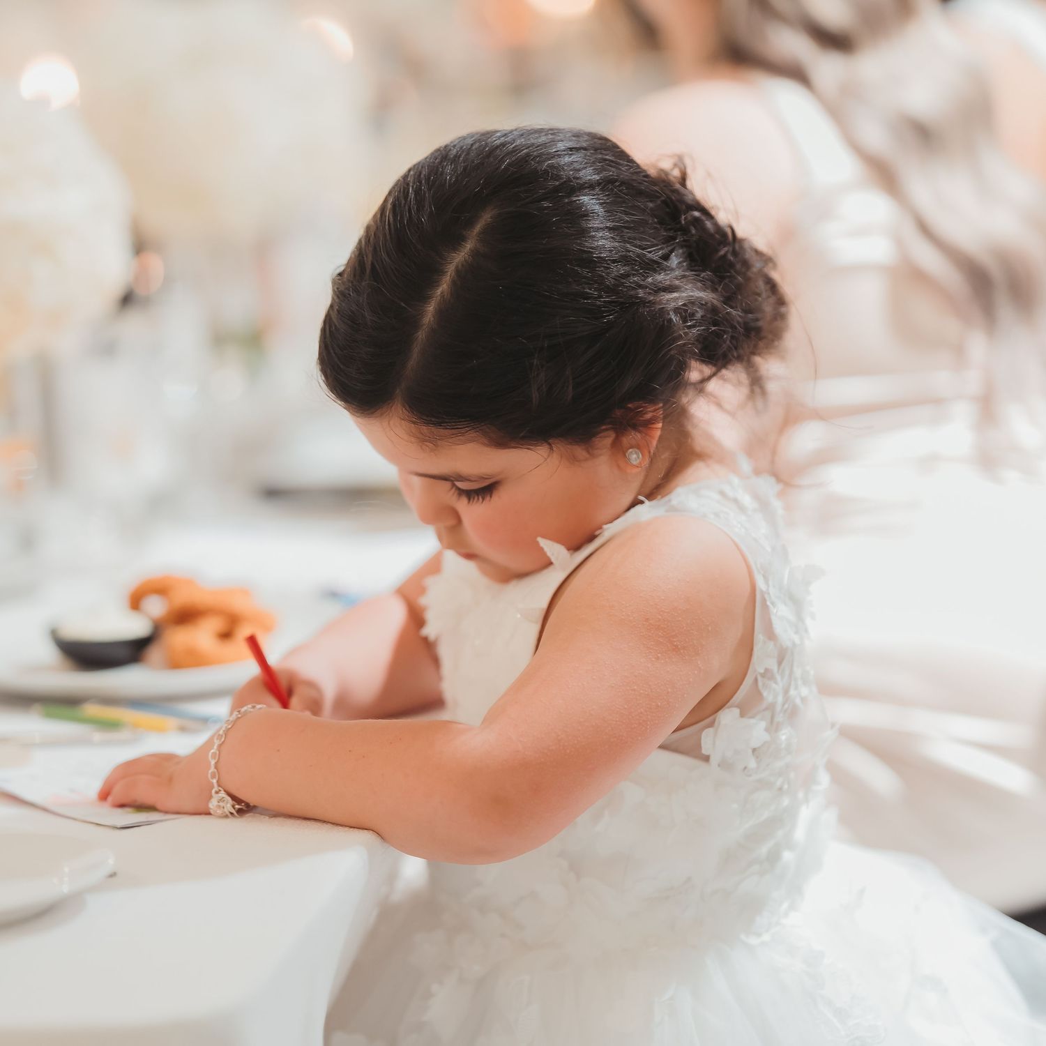 Young girl in a white dress writes or draws at a table during what appears to be a formal event.