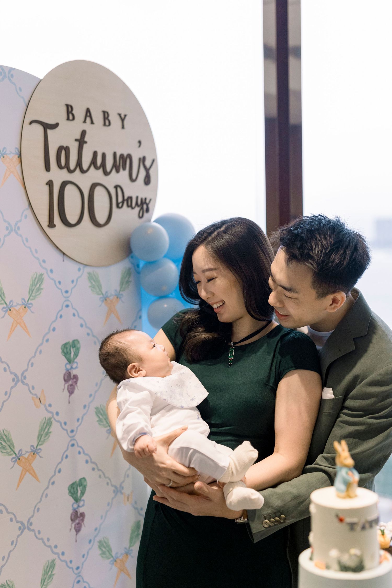A family celebrates their baby's 100 day milestone with balloons and decorative backdrop at a special party.
