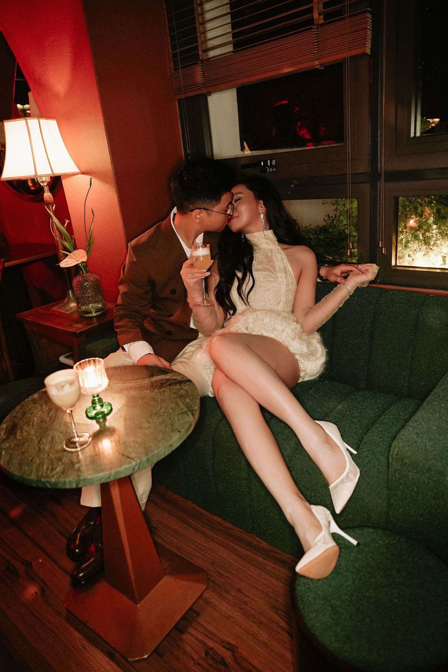 Romantic couple sharing a kiss on a green velvet sofa in a dimly lit bar with cocktails.