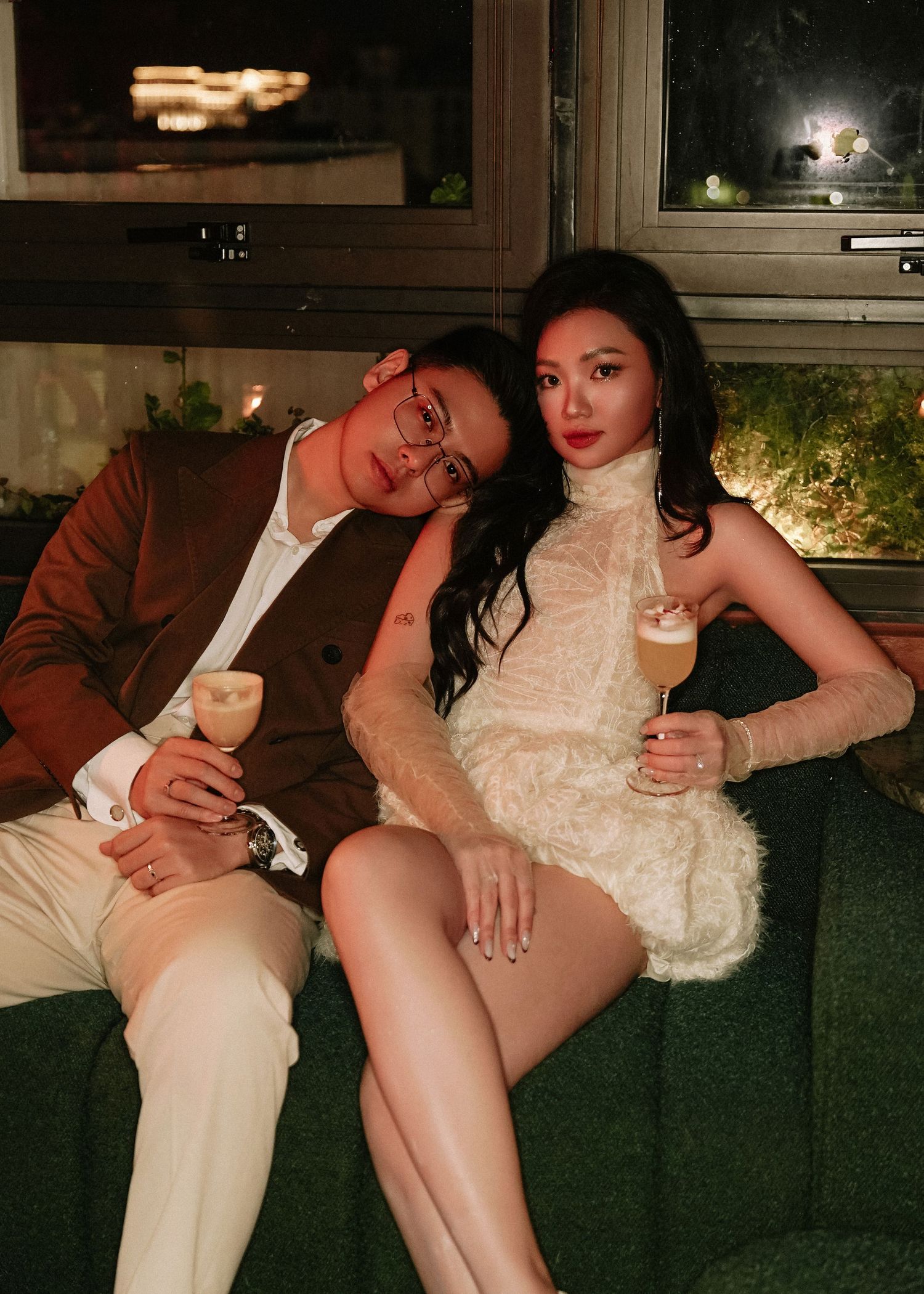 Stylish couple relaxing on a green sofa holding cocktails in a moody, warmly lit bar setting.