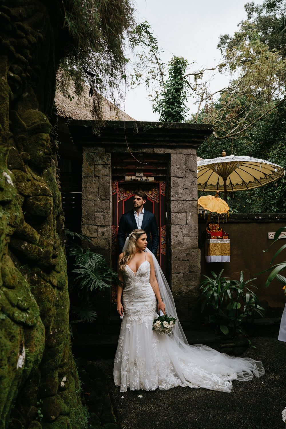 A wedding couple poses near a stone archway with decorative umbrellas in a garden setting.