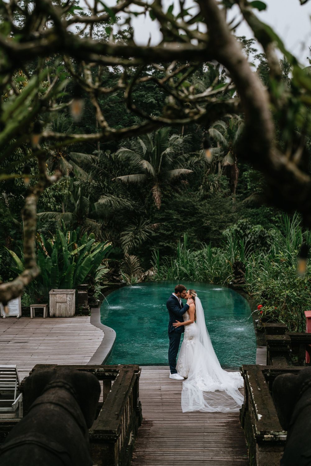 Wedding couple on wooden deck overlooking tropical infinity pool surrounded by greenery.